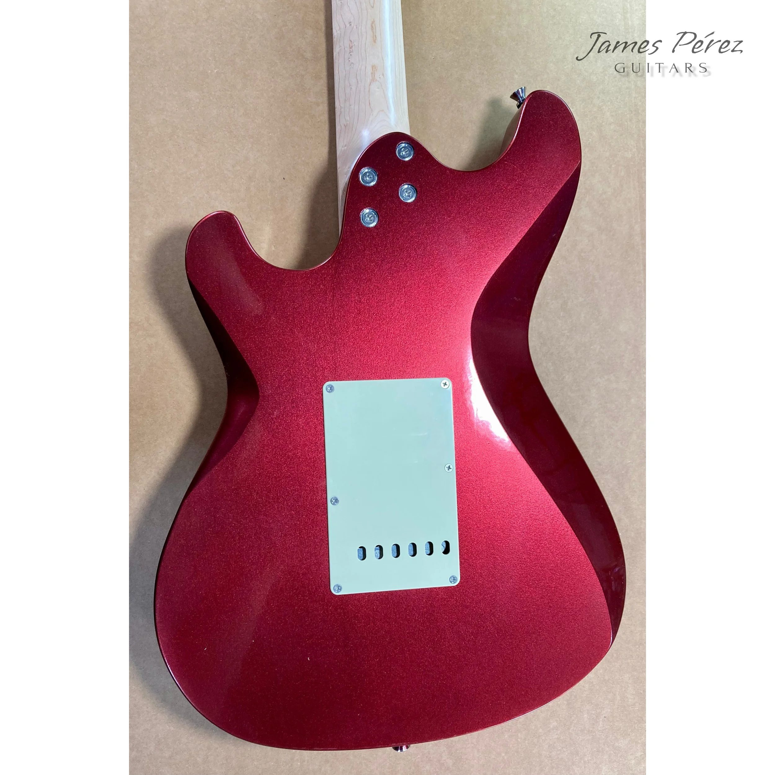 Back of a red electric guitar with a white cover plate, mounted on a beige background. The guitar has four control knobs and tuning pegs visible at the top. The top right corner has the words "James Pérez Guitars".