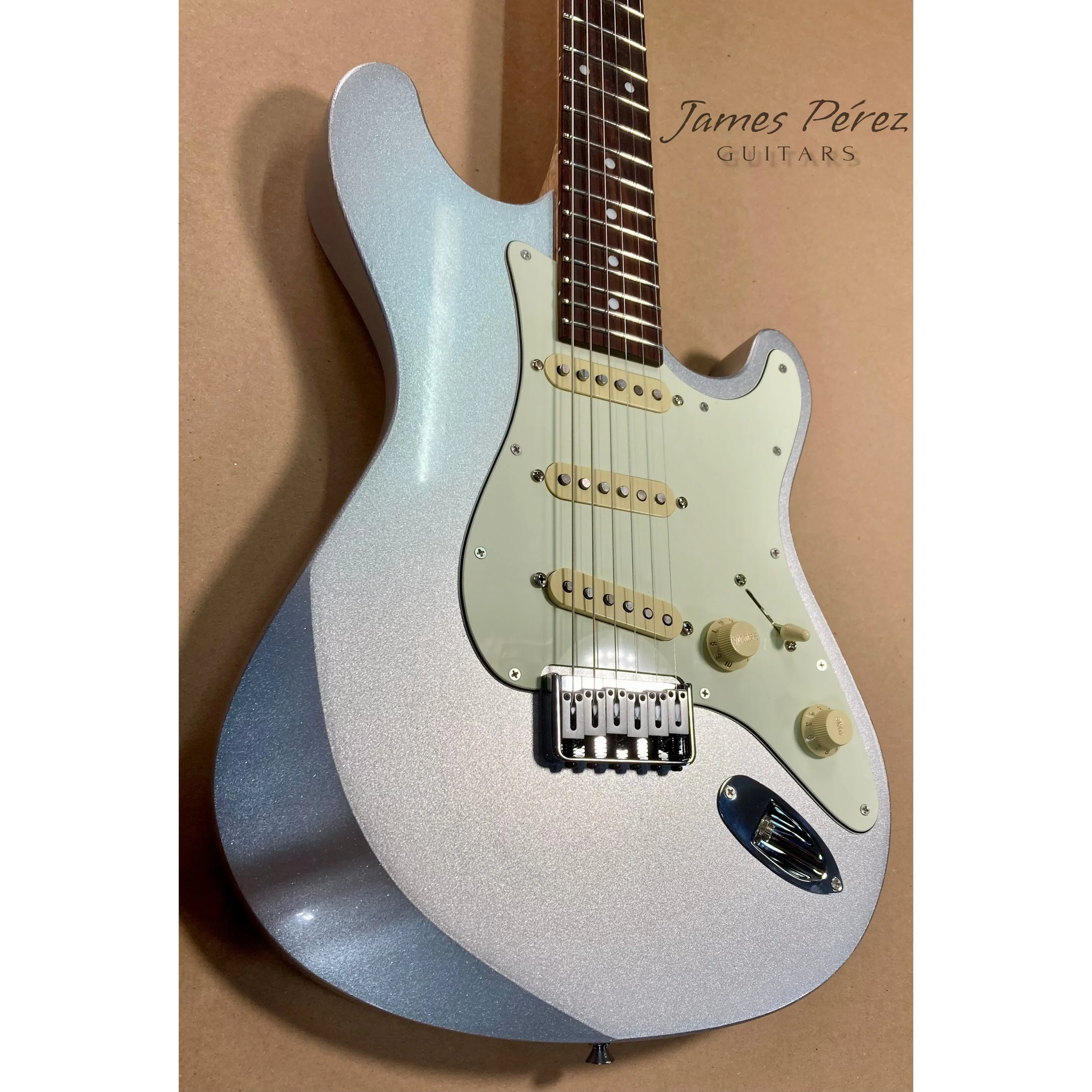 An electric guitar with a metallic silver finish, cream pickguard, three single-coil pickups, and control knobs, set against a brown background with the text "James Pérez GUITARS" in the top right corner.