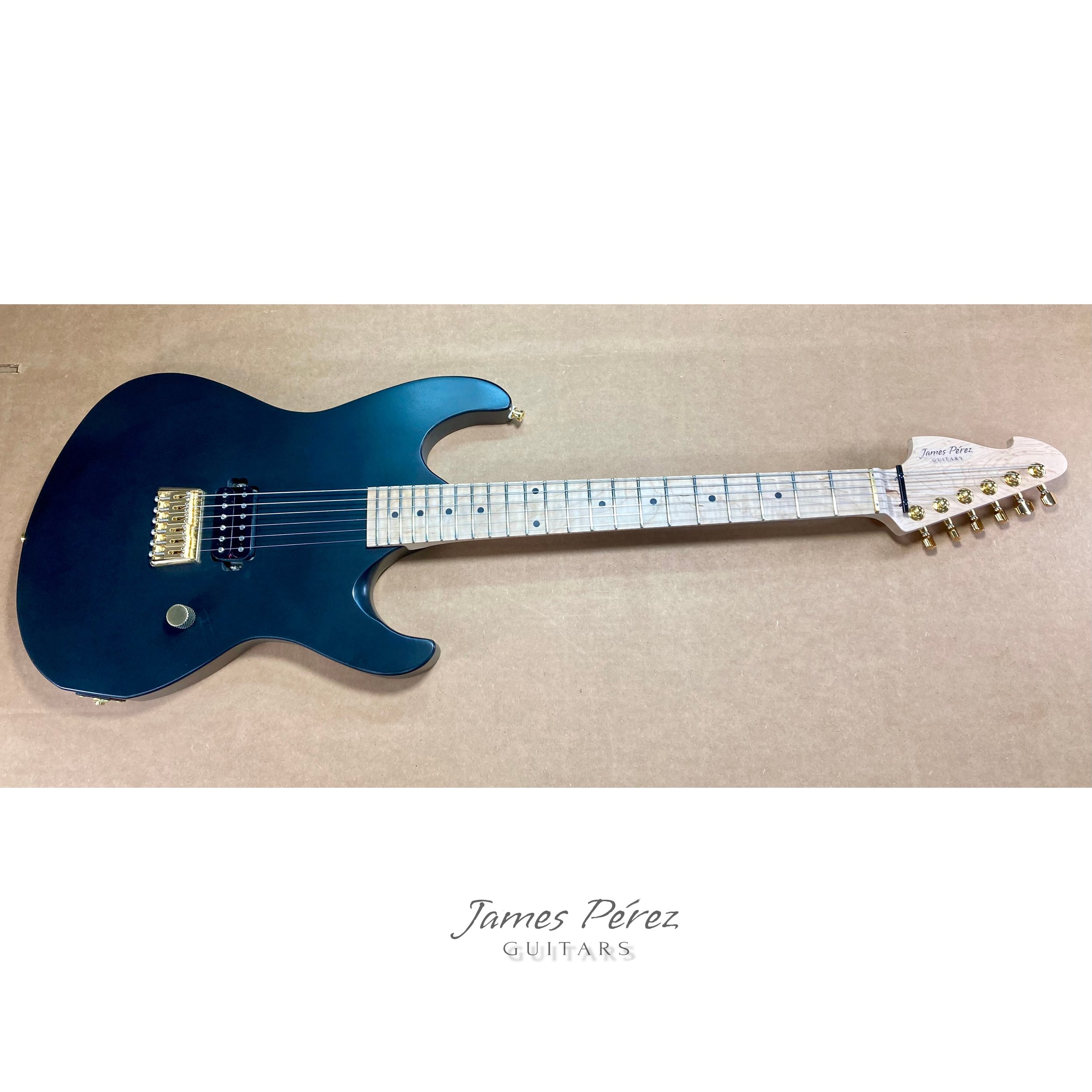 Electric guitar with a black body, white fretboard, and gold tuning pegs, resting on a beige surface. Text on the image reads "James Pérez Guitars."