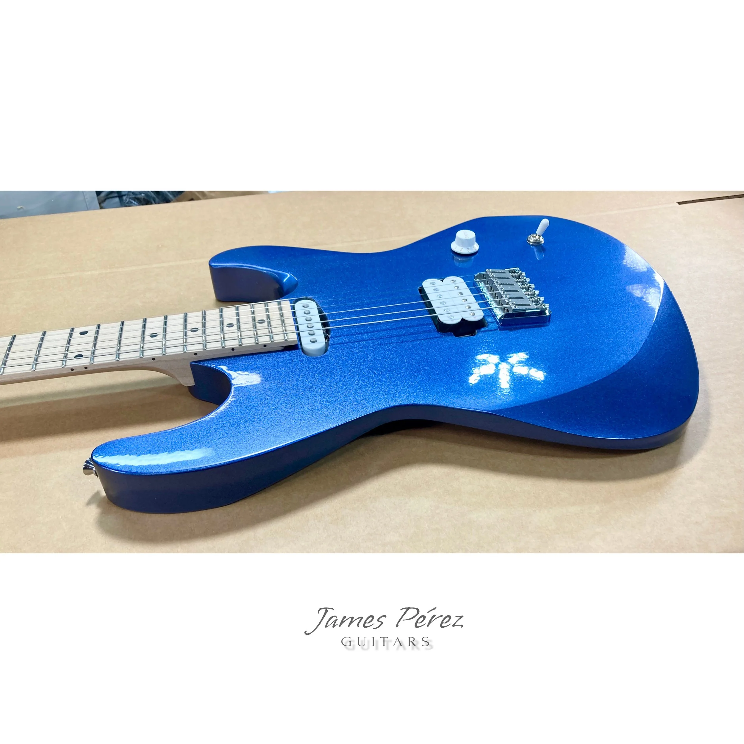 Blue electric guitar lying on a cardboard surface with a white label reading 'James Pérez GUITARS' underneath.