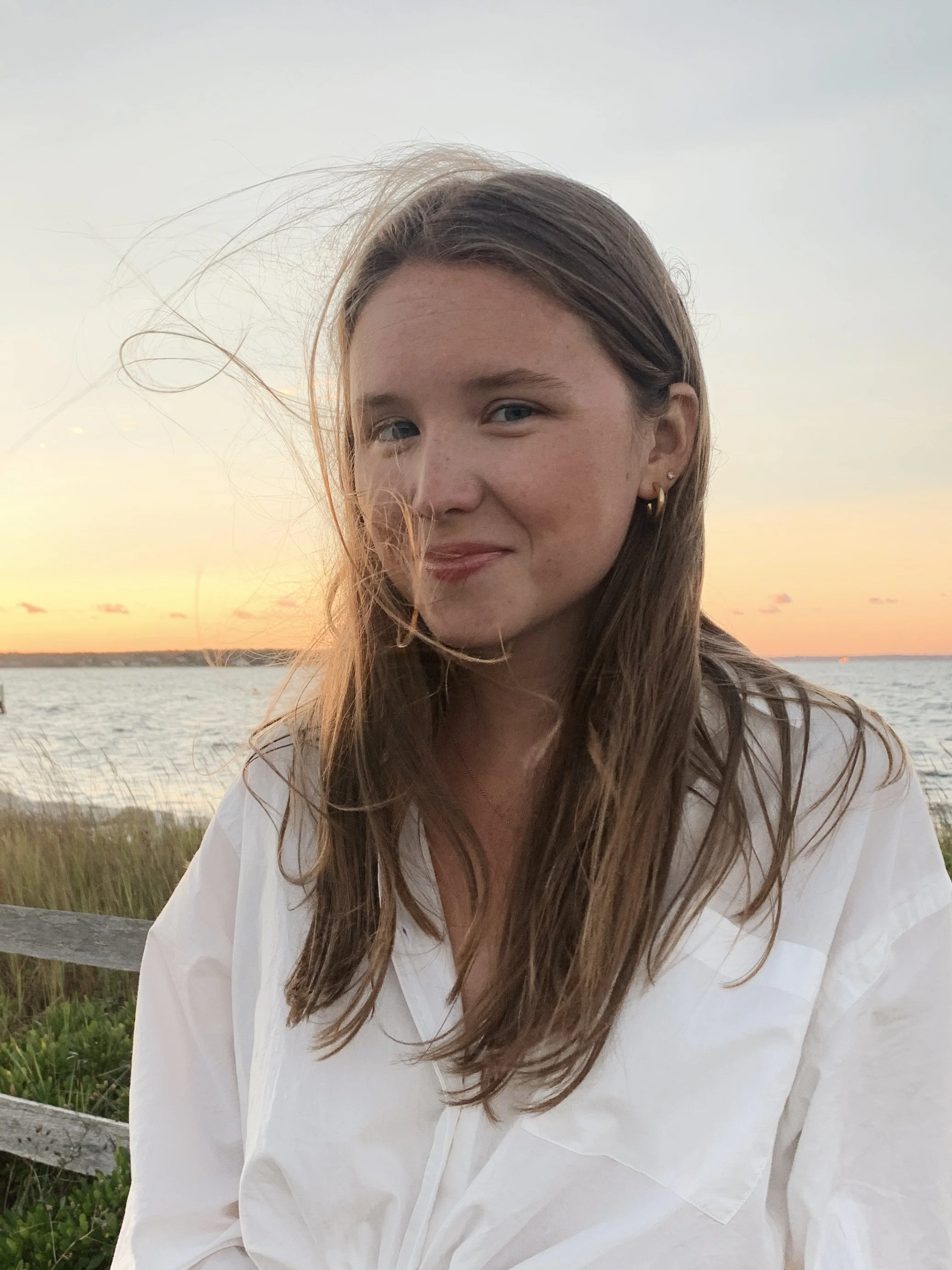 A young woman with long brown hair, wearing a white shirt, standing outdoors near a body of water during sunset.