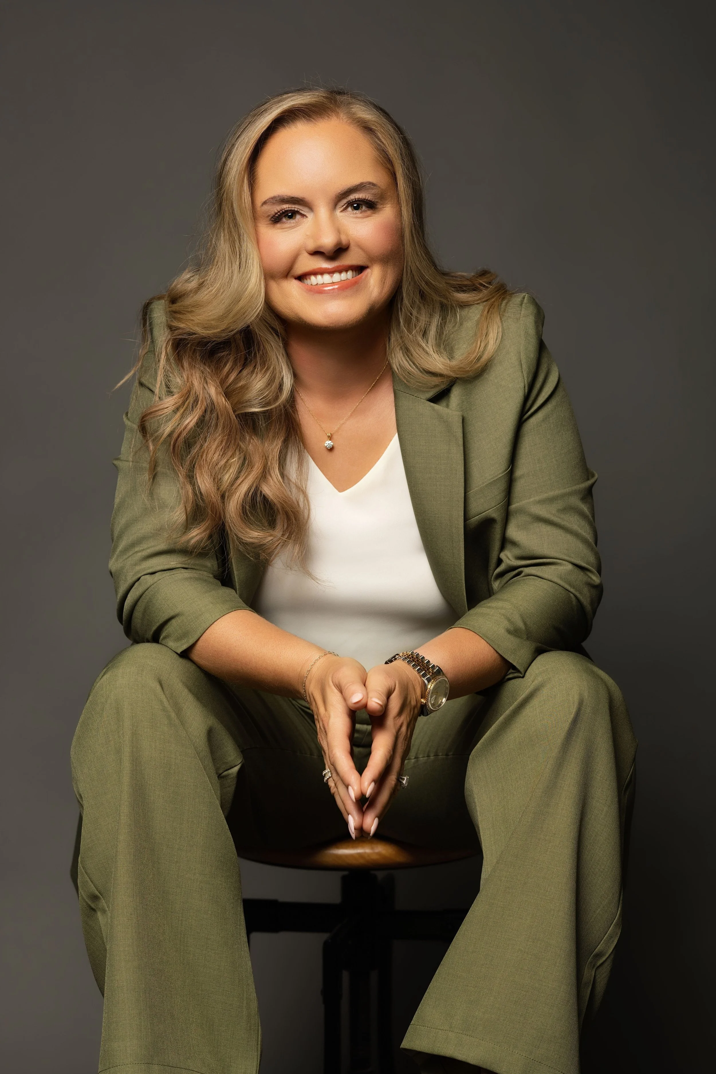 A woman with long wavy blonde hair smiling, wearing a green suit and white top, sitting on a stool against a grey background.