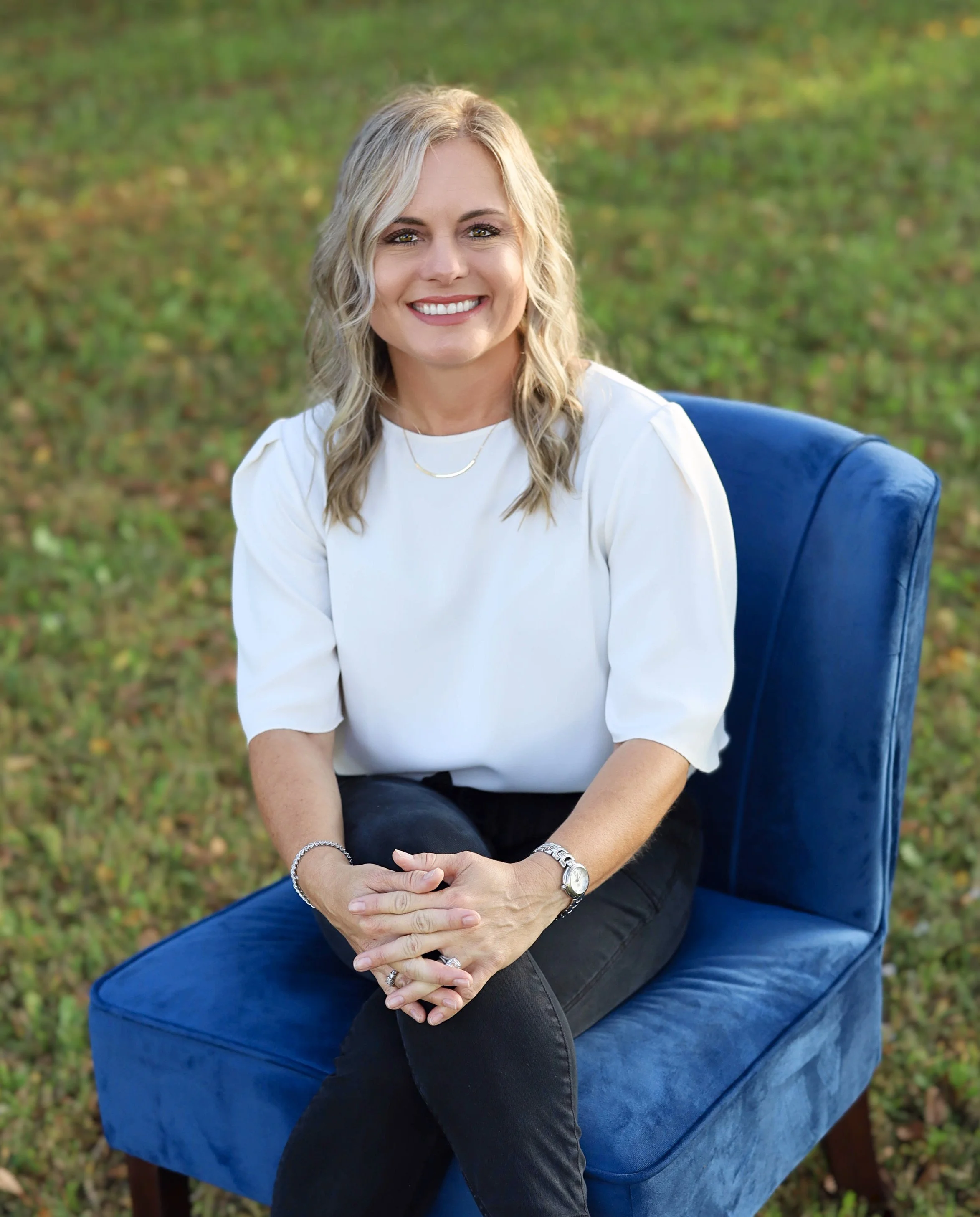 A woman with blonde hair smiling, sitting on a blue velvet chair outdoors on grass. She is wearing a white top, black pants, a watch, and jewelry.
