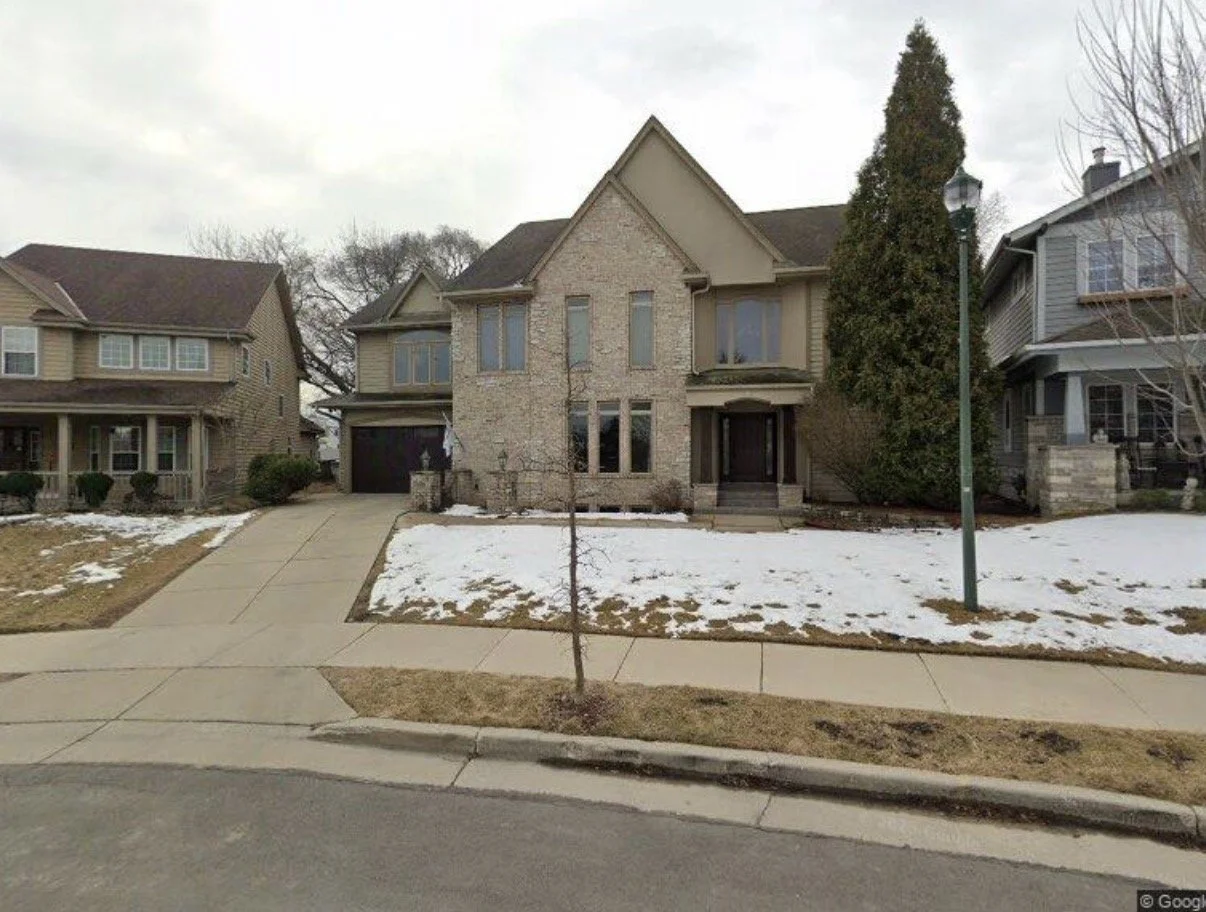 A suburban neighborhood with three houses, a sidewalk, a small tree, and a streetlamp. The houses have brick and siding exteriors, with some snow on the lawn.