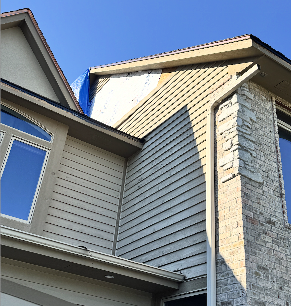 Close-up of a house under construction with new siding, brick chimney, and partially installed roof covering, against a clear blue sky.