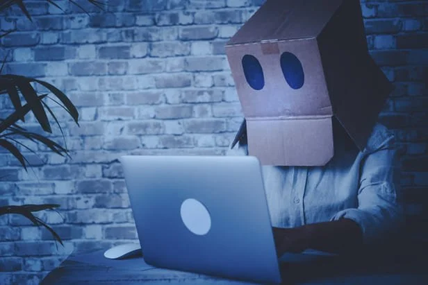 Pessoa com uma caixa de papelão na cabeça diante de um laptop, ilustrando o chatbot engessado no atendimento de clínicas.