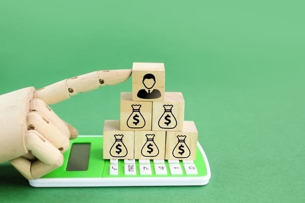 Mão de madeira empilhando blocos com cifras e um paciente no topo sobre uma calculadora, representando o impacto financeiro do agente de IA na agenda da clínica.
