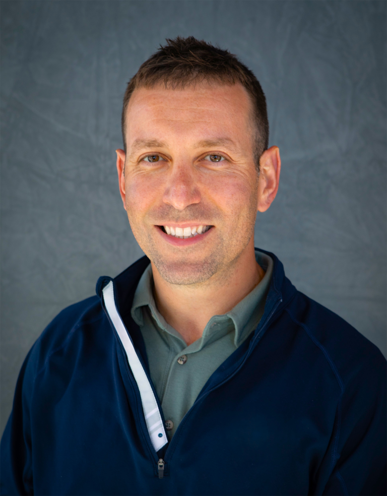 A smiling young man with short brown hair wearing a gray collared shirt and a navy blue jacket posing in front of a gray background.