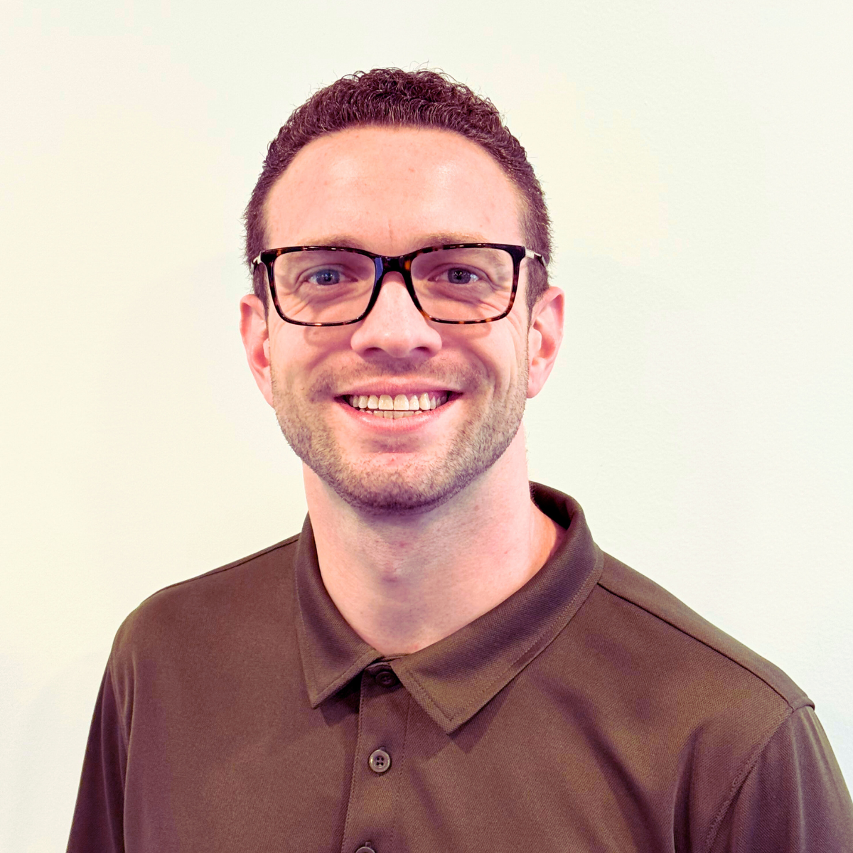 Portrait of a smiling man with short dark hair, glasses, wearing a brown polo shirt, against a plain light background.