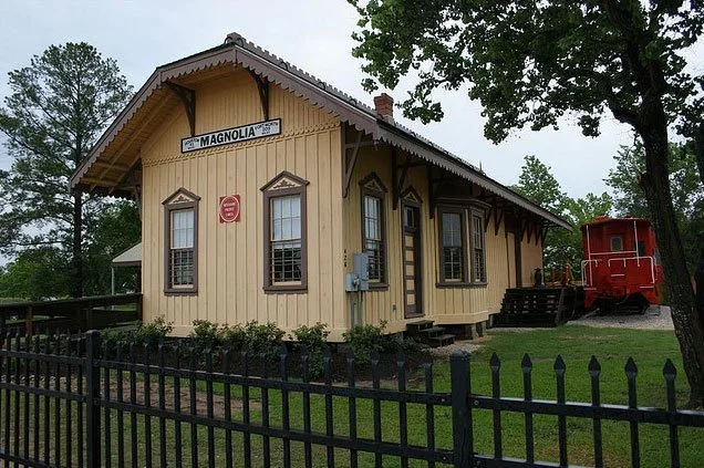 Historic train station labeled 'Magnolia,' made of wood with decorative trim, surrounded by a black fence and trees, with a red caboose nearby.