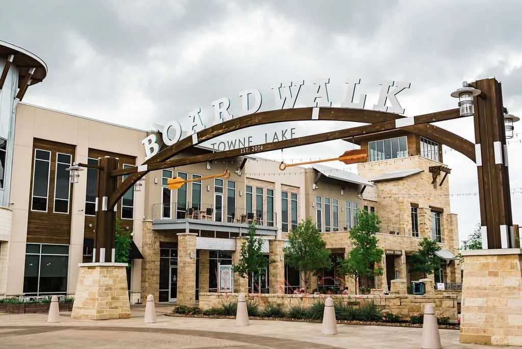The entrance of Boward Walk at Towne Lake, with a large wooden archway sign and decorative oar, surrounded by modern buildings and trees.