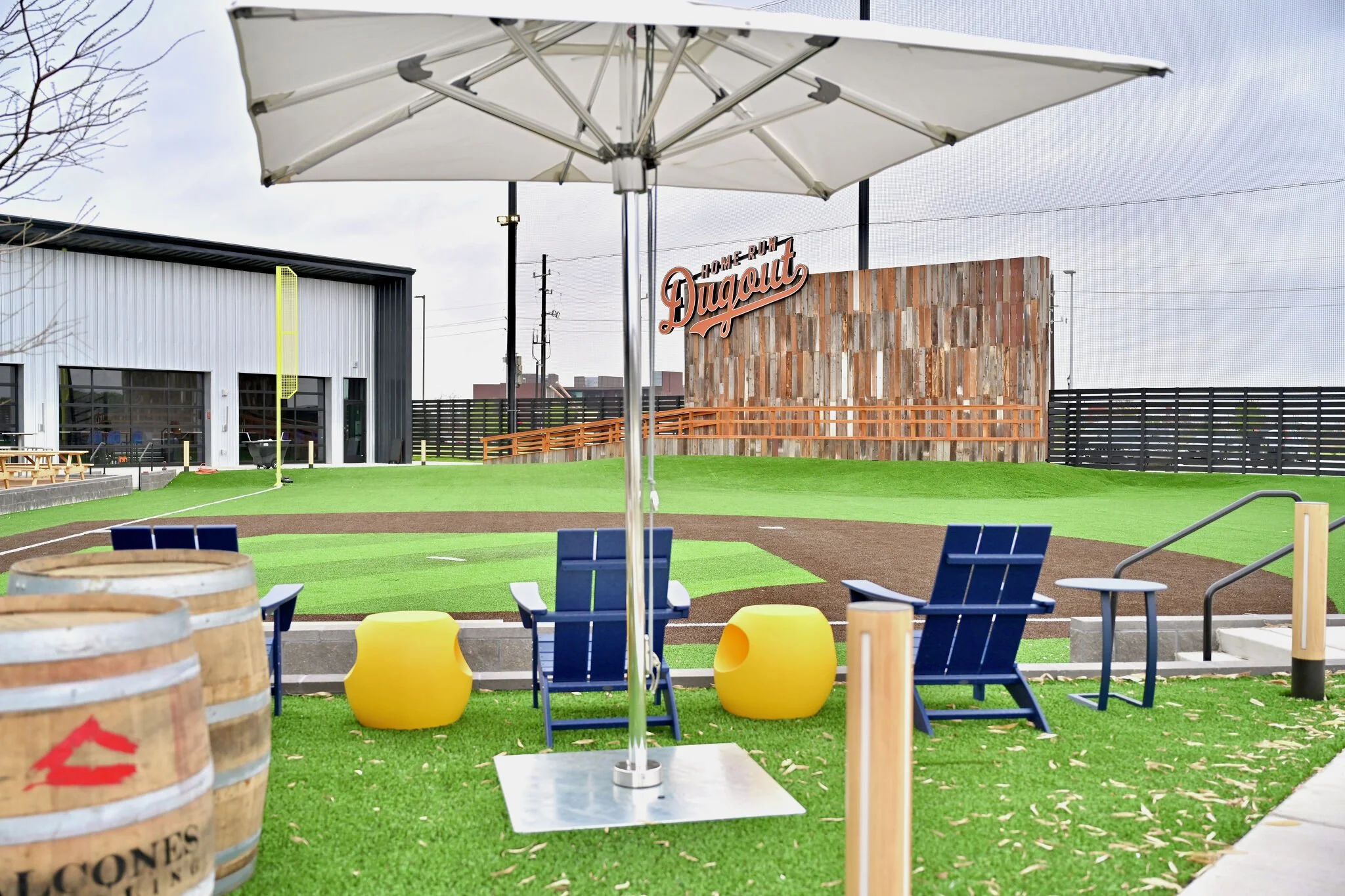 Outdoor patio area with chairs, yellow stools, and a large white umbrella, overlooking a baseball field with a Dugout sign and a building in the background.