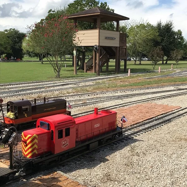 A small red model train on tracks in a park, with a wooden observation tower and trees in the background.