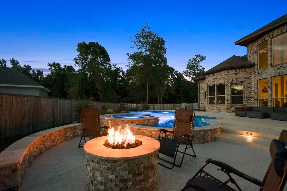 Backyard with a lit fire pit, swimming pool, patio seating, and outdoor lighting at dusk.