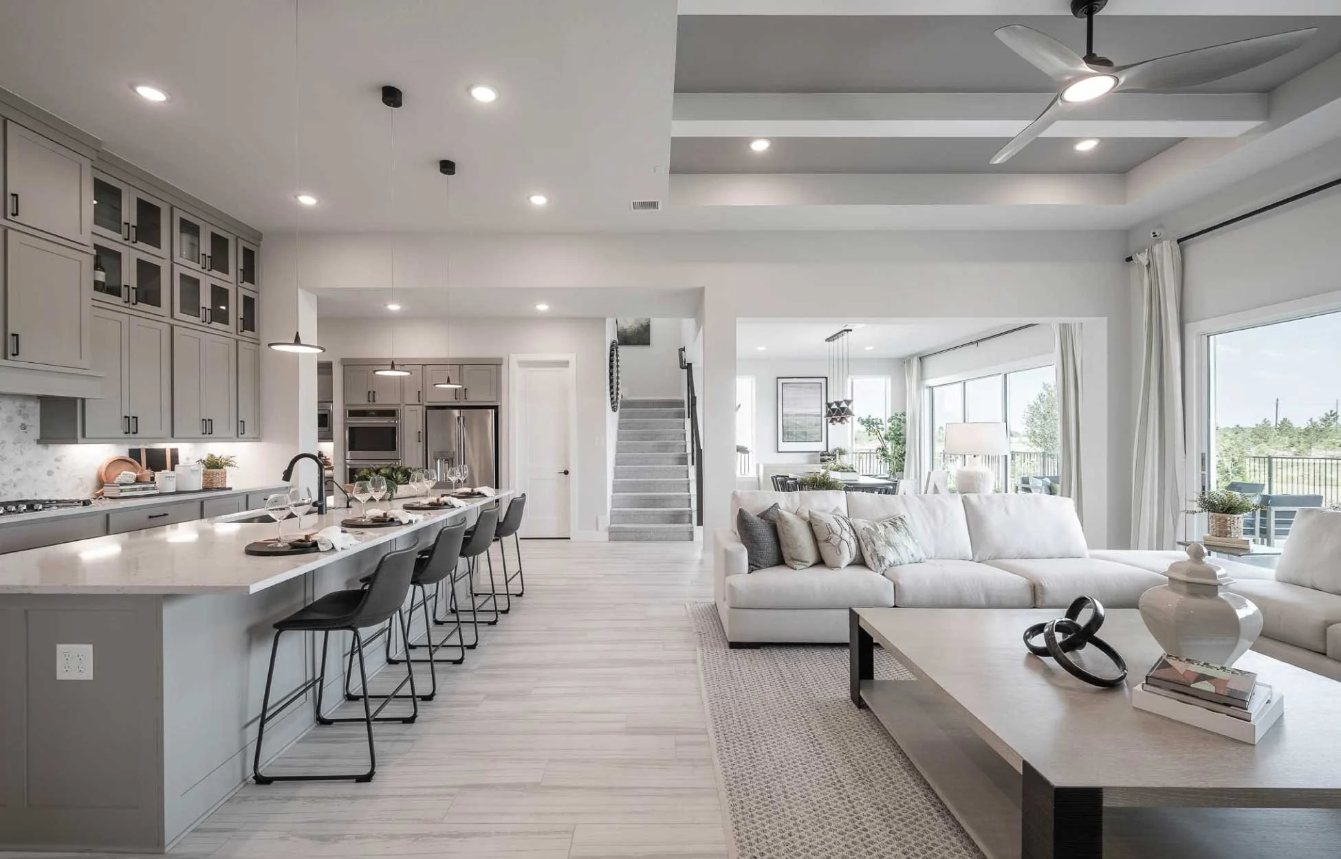 Open-concept living room and kitchen with gray cabinetry, island with bar stools, white sofas, large windows, and ceiling fan.