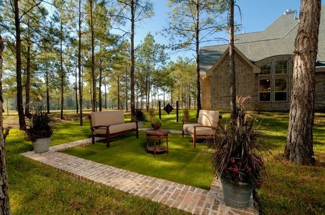 Outdoor backyard patio with two cream-colored chairs, a small round table, and potted plants on brick and grass surface, surrounded by trees and a brick house in the background.