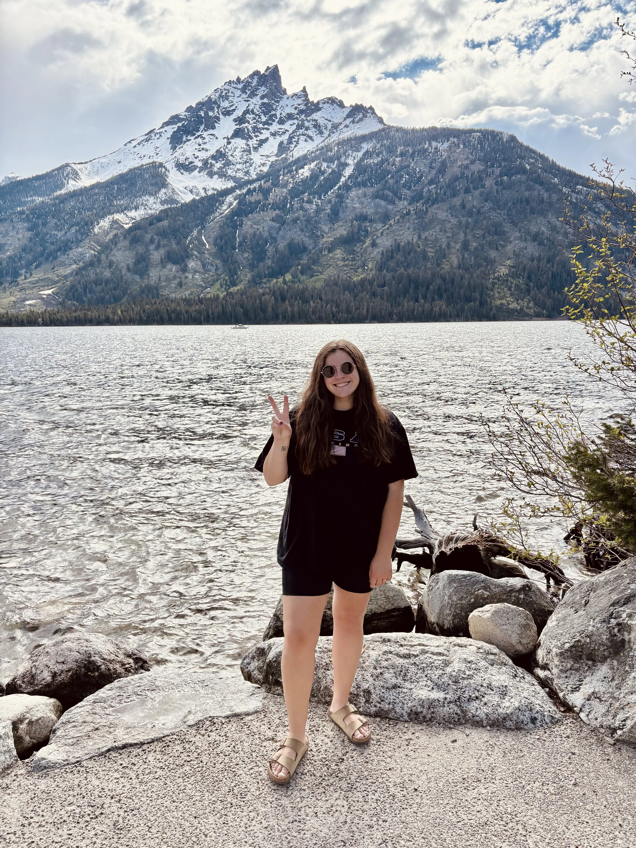 A young woman with long brown hair, wearing sunglasses, a black T-shirt, black shorts, and sandals, standing on a rocky lakeshore making a peace sign with her right hand, with a large mountain with snow at its peak and a partly cloudy sky in the background.