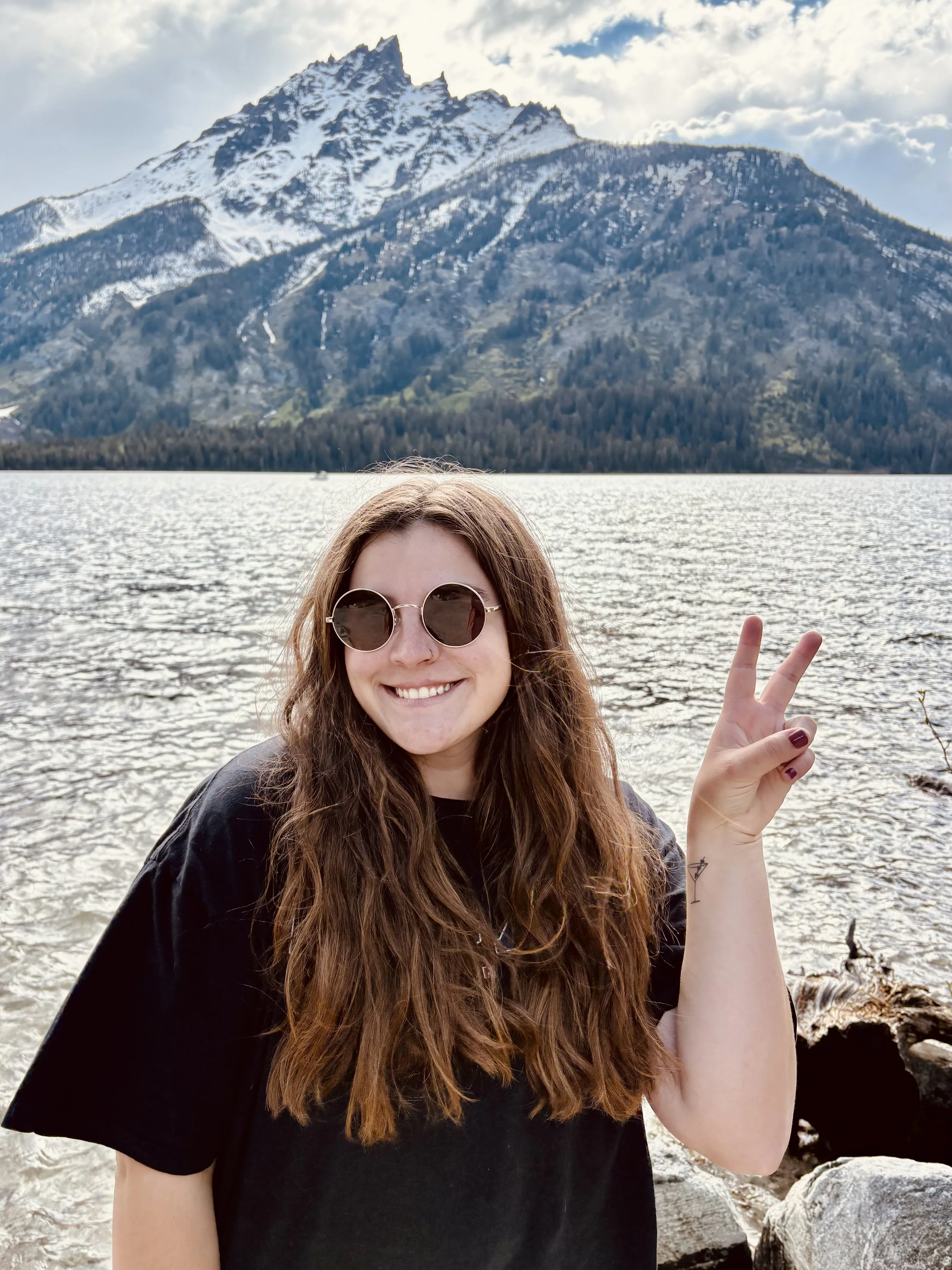 A young woman with long brown hair, wearing sunglasses and a black shirt, smiling and making a peace sign with her right hand in front of a lake with mountains in the background.