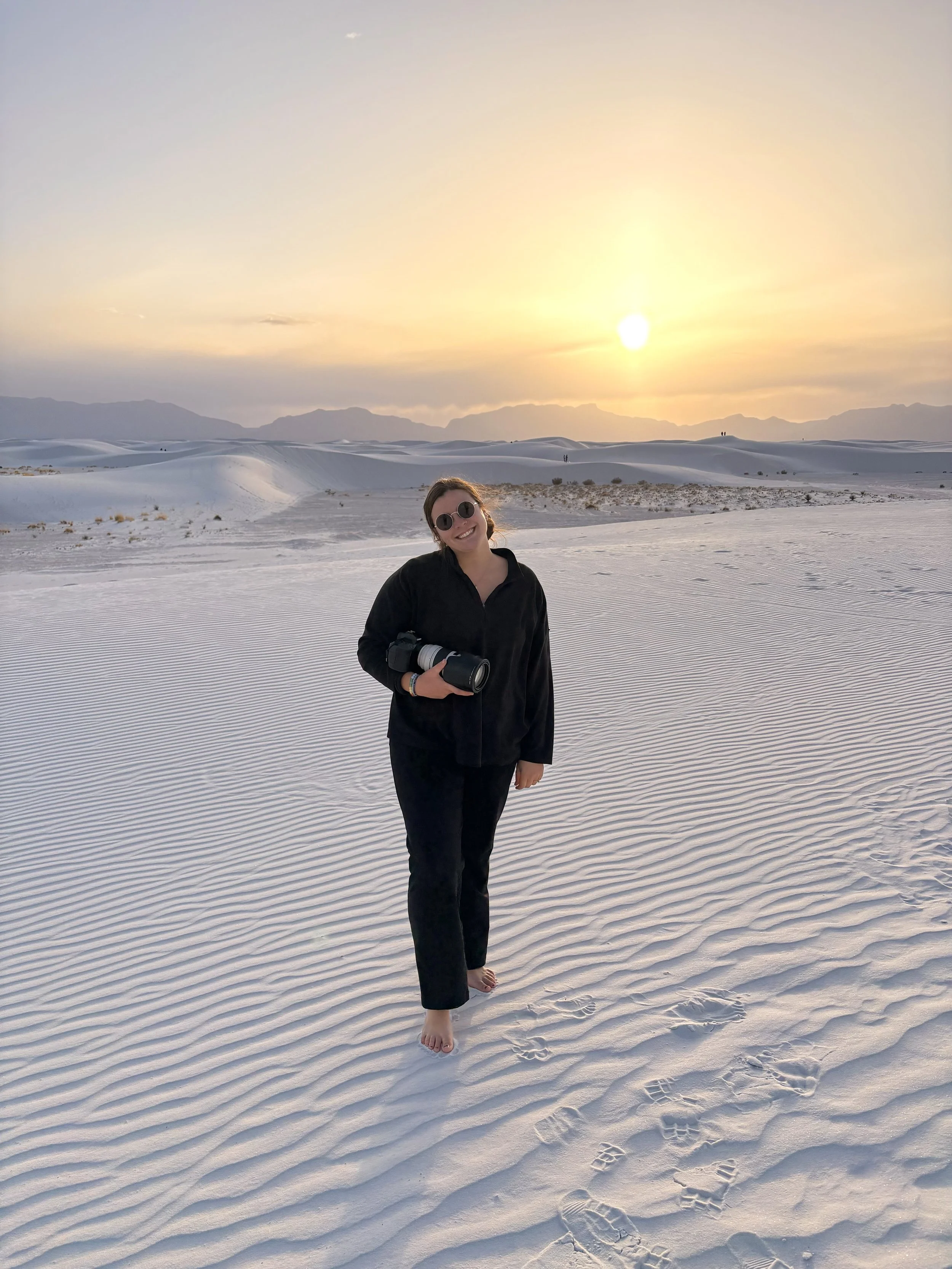 A woman smiling in black clothing and sunglasses holding a camera, standing barefoot in white sand dunes at sunset.
