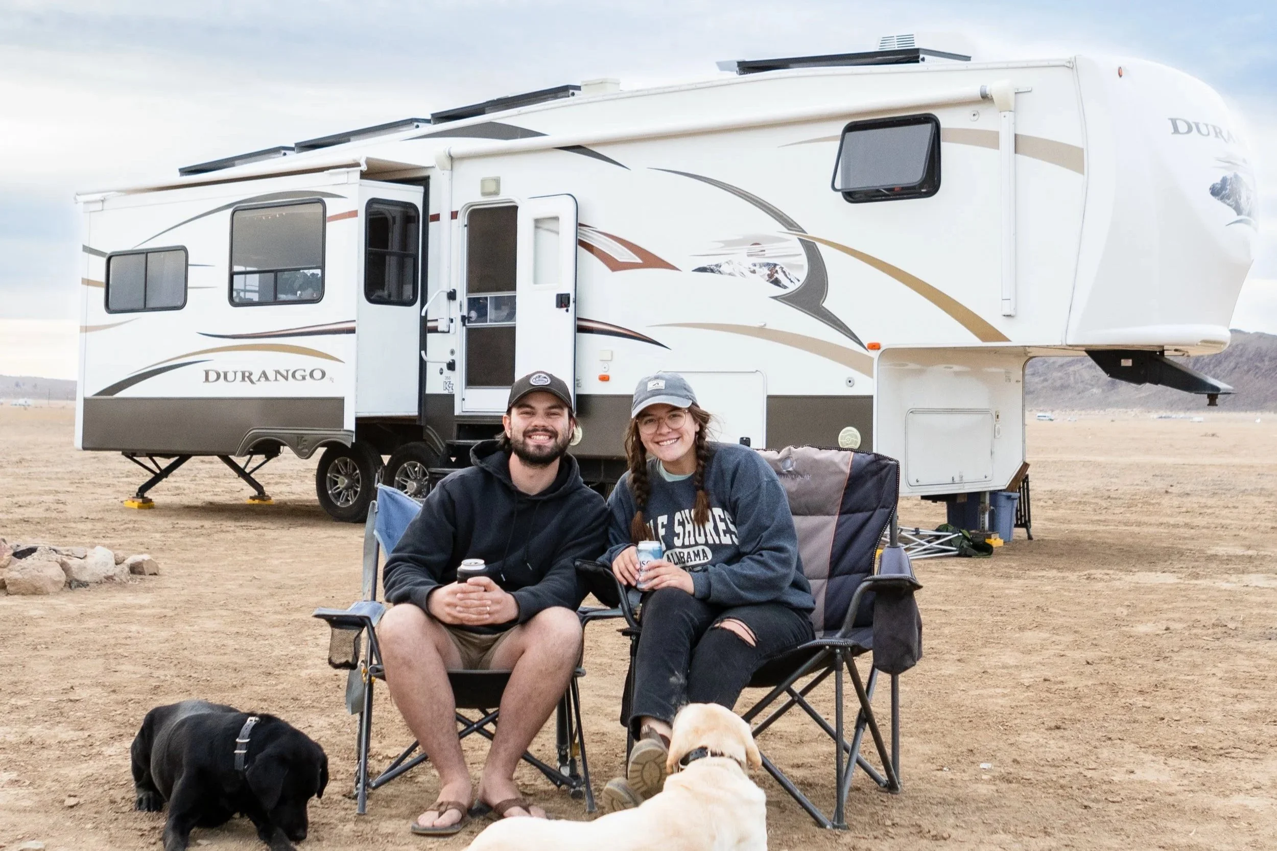 Young couple sitting in camping chairs with two dogs, in front of a large white Durango fifth wheel RV, on a barren landscape with mountains in the background.