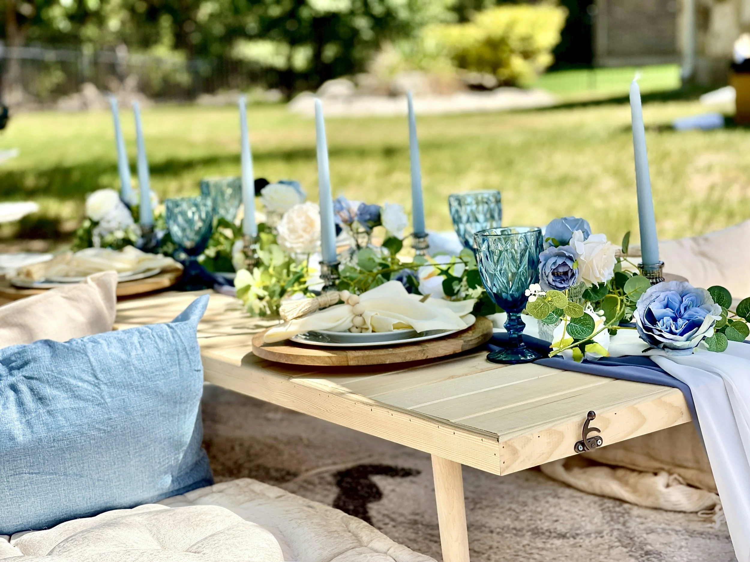 Outdoor table set for a special occasion with blue and white flowers, blue glassware, white candles, and elegant tableware, surrounded by cushions on the ground.