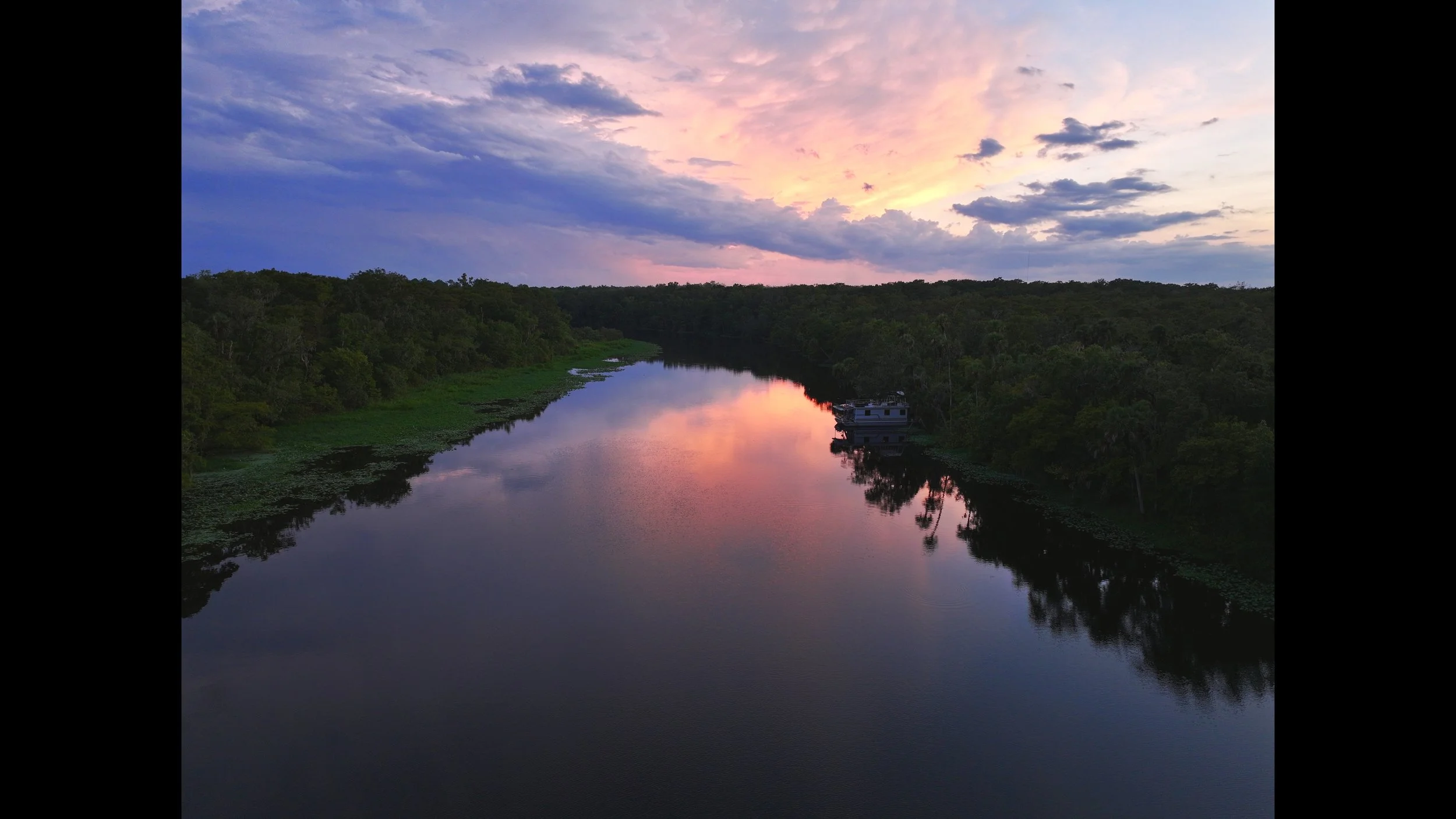 A serene river at sunset with colorful sky and clouds reflecting on the water. Trees line both sides of the river, with a houseboat partially visible on the right bank. St. John's River Florida. DJI Mavic 3 Pro. Professional drone photography.