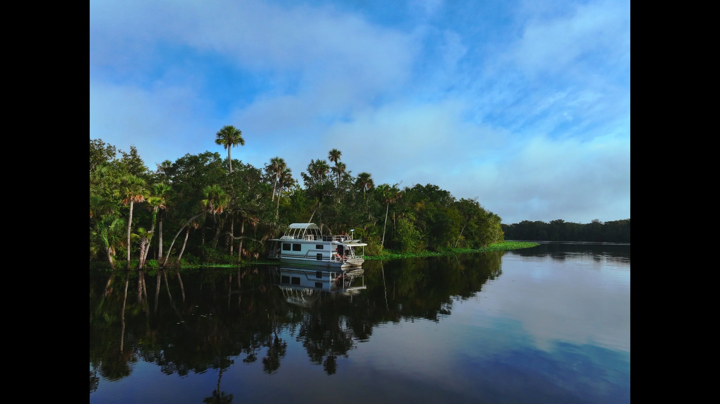 A houseboat anchored along a calm St. John's River, surrounded by dense tropical trees and palm trees, with a partly cloudy blue sky reflected in the water.  DeLand, Florida. DJI Mavic 3 Pro. Professional drone photography.