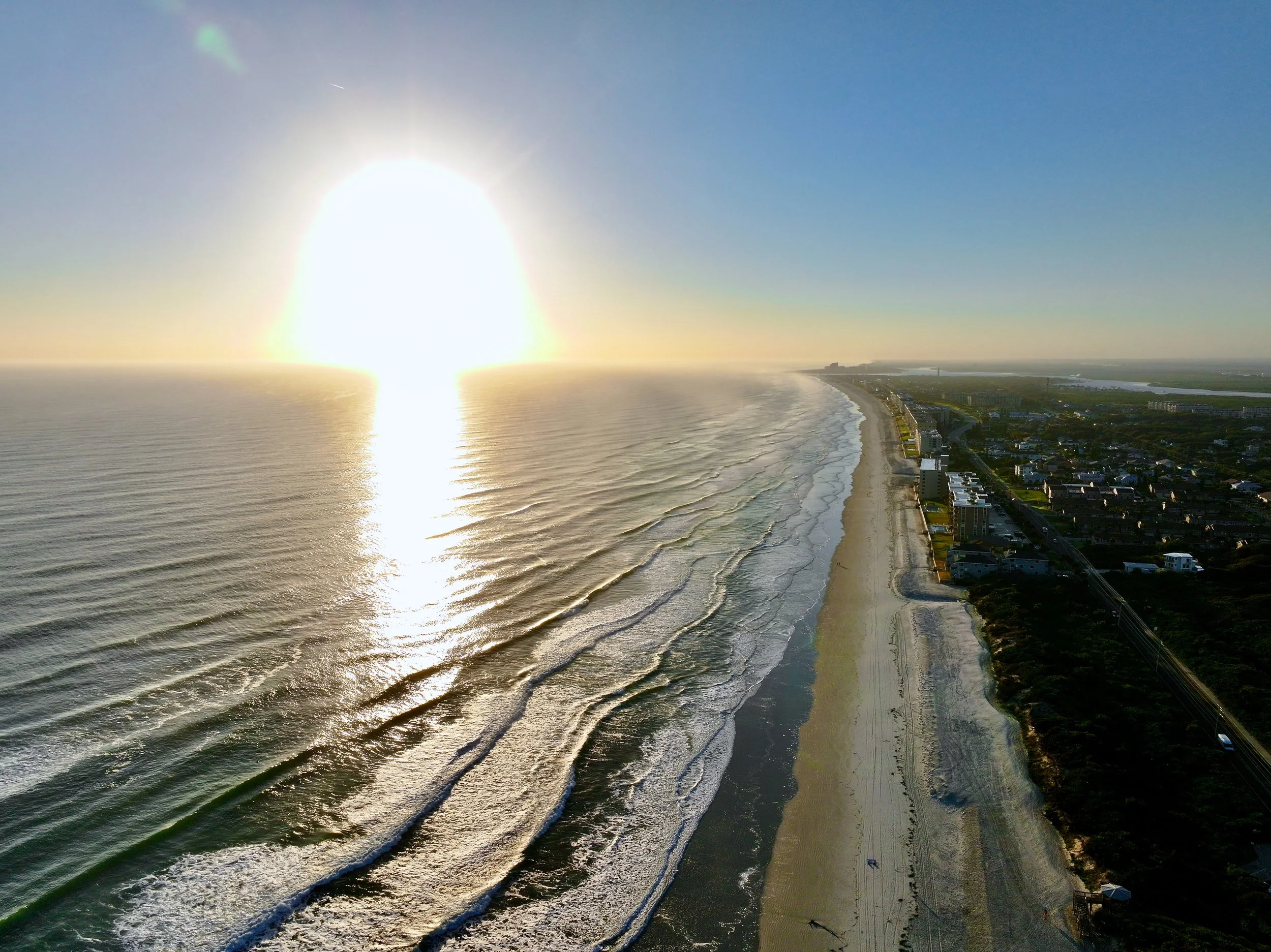 An aerial view of a beach at sunset with ocean waves, a shoreline, and buildings along the coast.