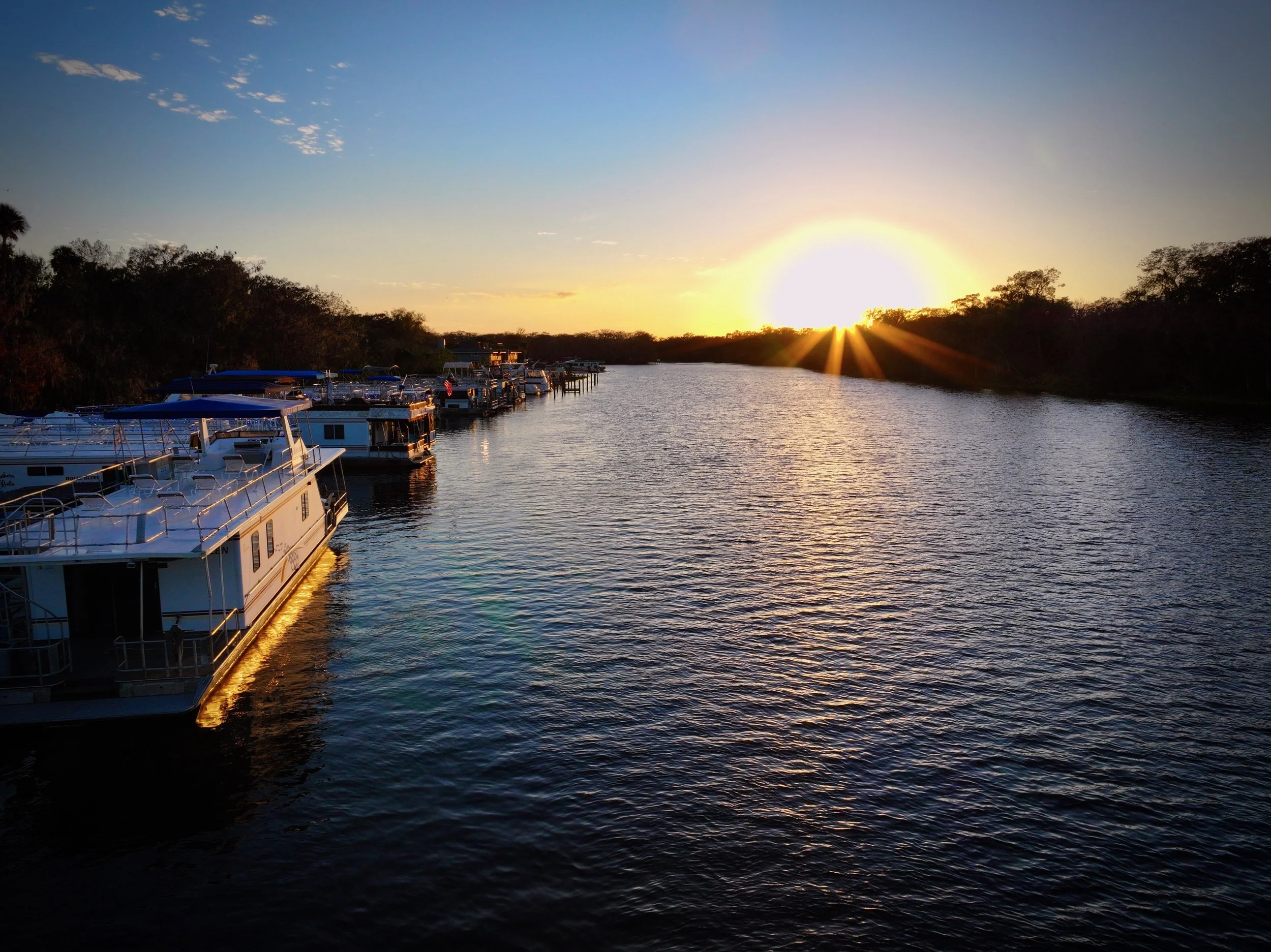 A row of boats docked along the St. John's River at sunset with the sun near the horizon, casting golden light on the water and silhouettes of trees in the background.  DeLand, Florida.  DJI Mavic 3 Pro.  Professional drone photography.