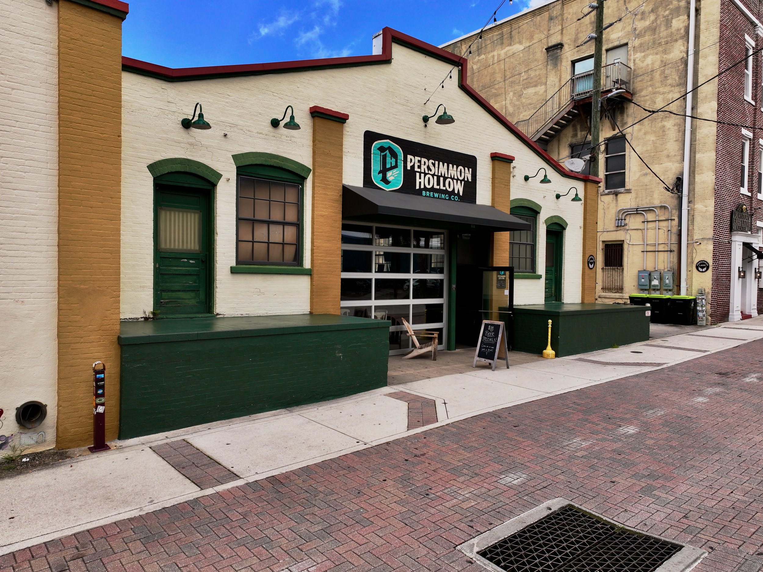 Exterior view of Persimmon Hollow Brewing Co. with a black sign, green door, green window frames. Brick sidewalk and neighboring buildings are visible.  Artisan Alley, DeLand, Florida.  DJI Mavic 3 Pro.  Professional drone photography.