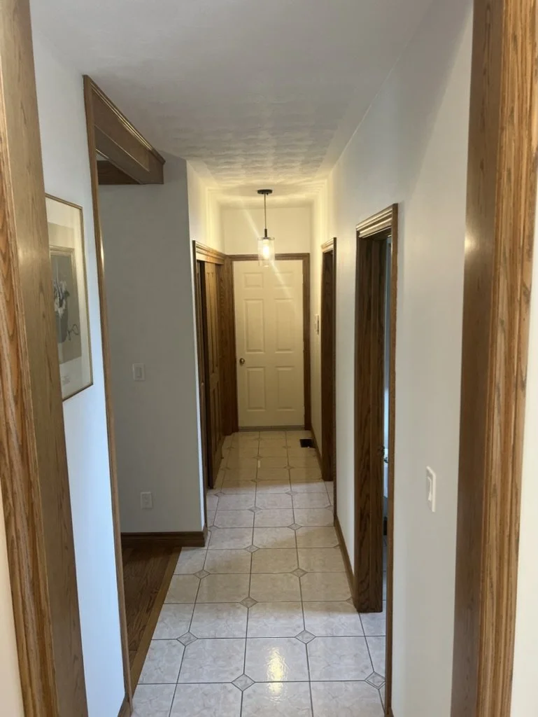 View of a hallway with tiled flooring, white walls, wooden trim, and a white door at the end with a hanging ceiling light.