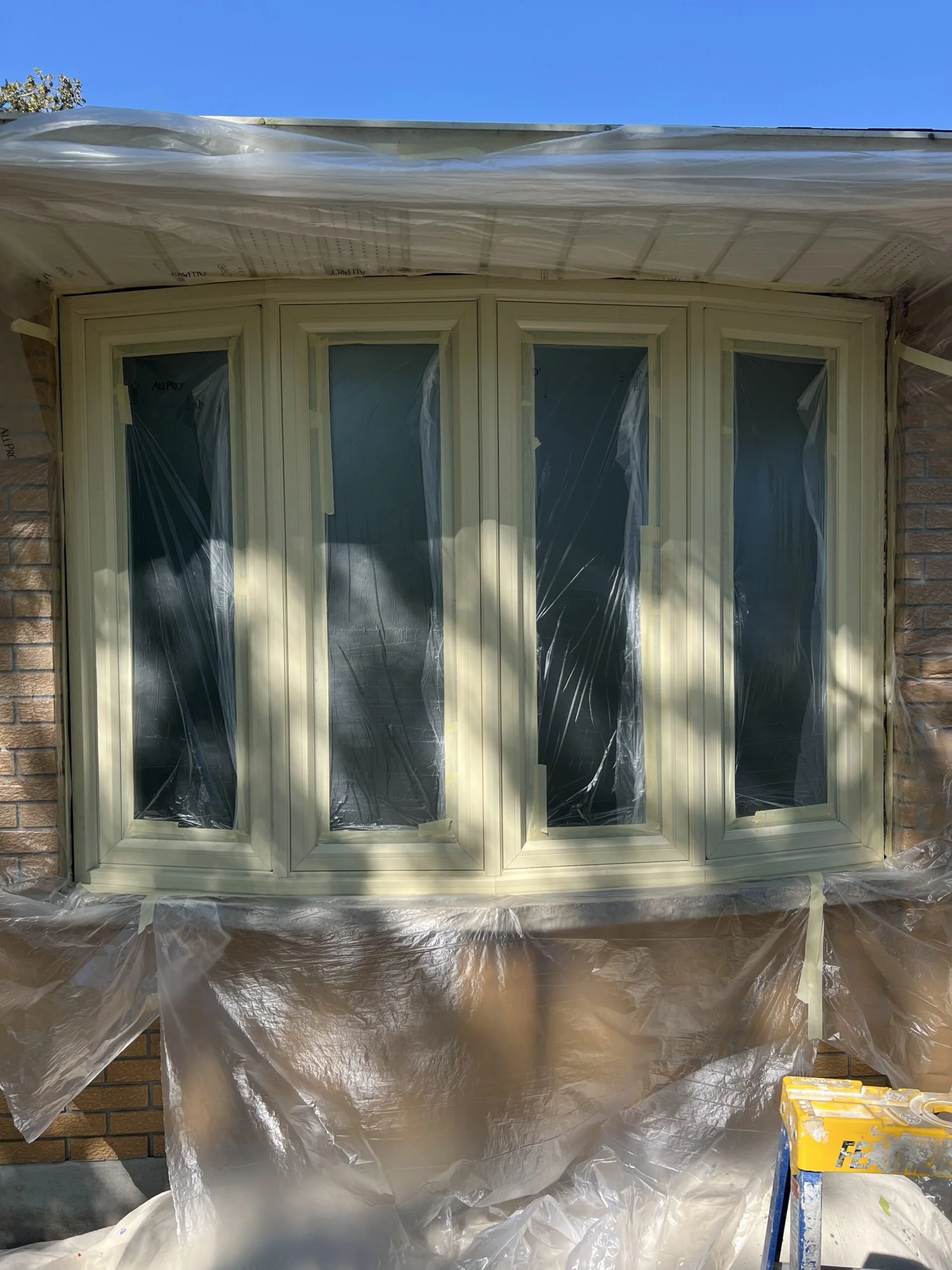 Brick house with a newly installed bay window covered in plastic and painter's tape, surrounded by plastic sheeting for painting or renovation work.