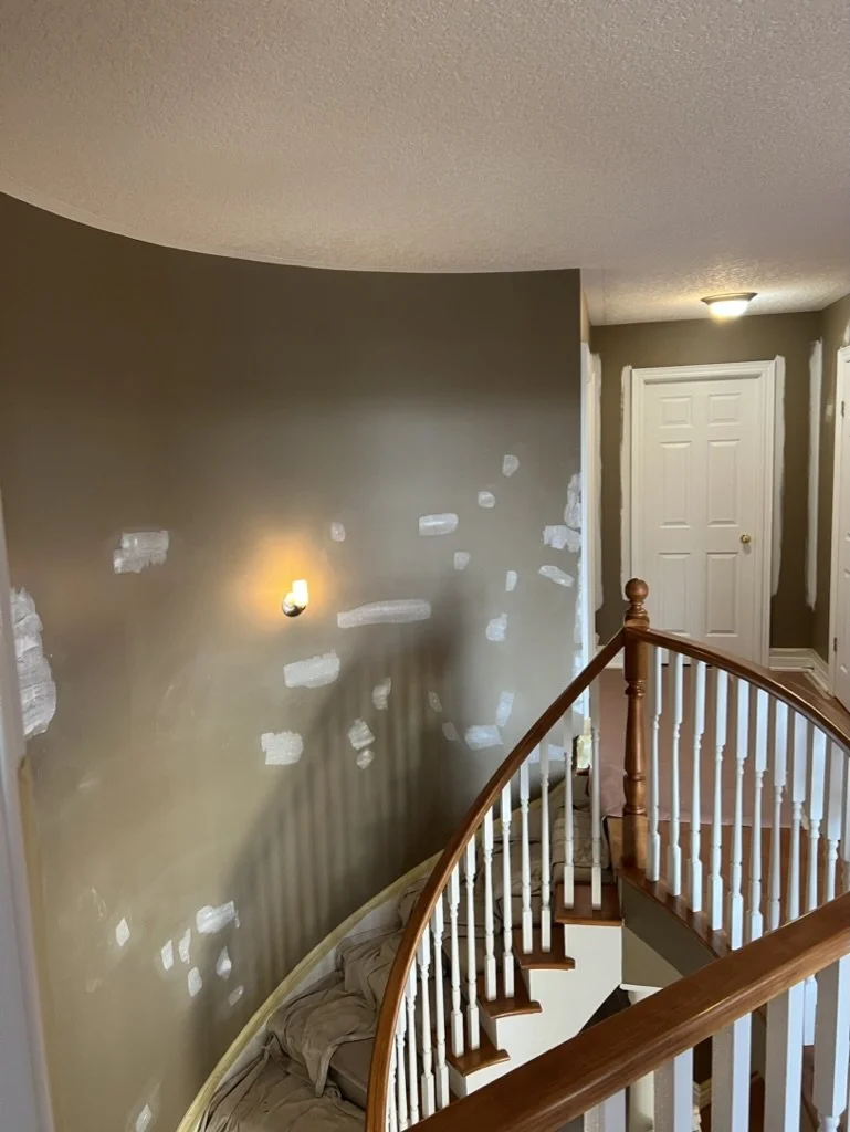 Interior of a staircase landing area under renovation, with drywall patches on the wall, a wooden railing, a covered couch, and closed white door.