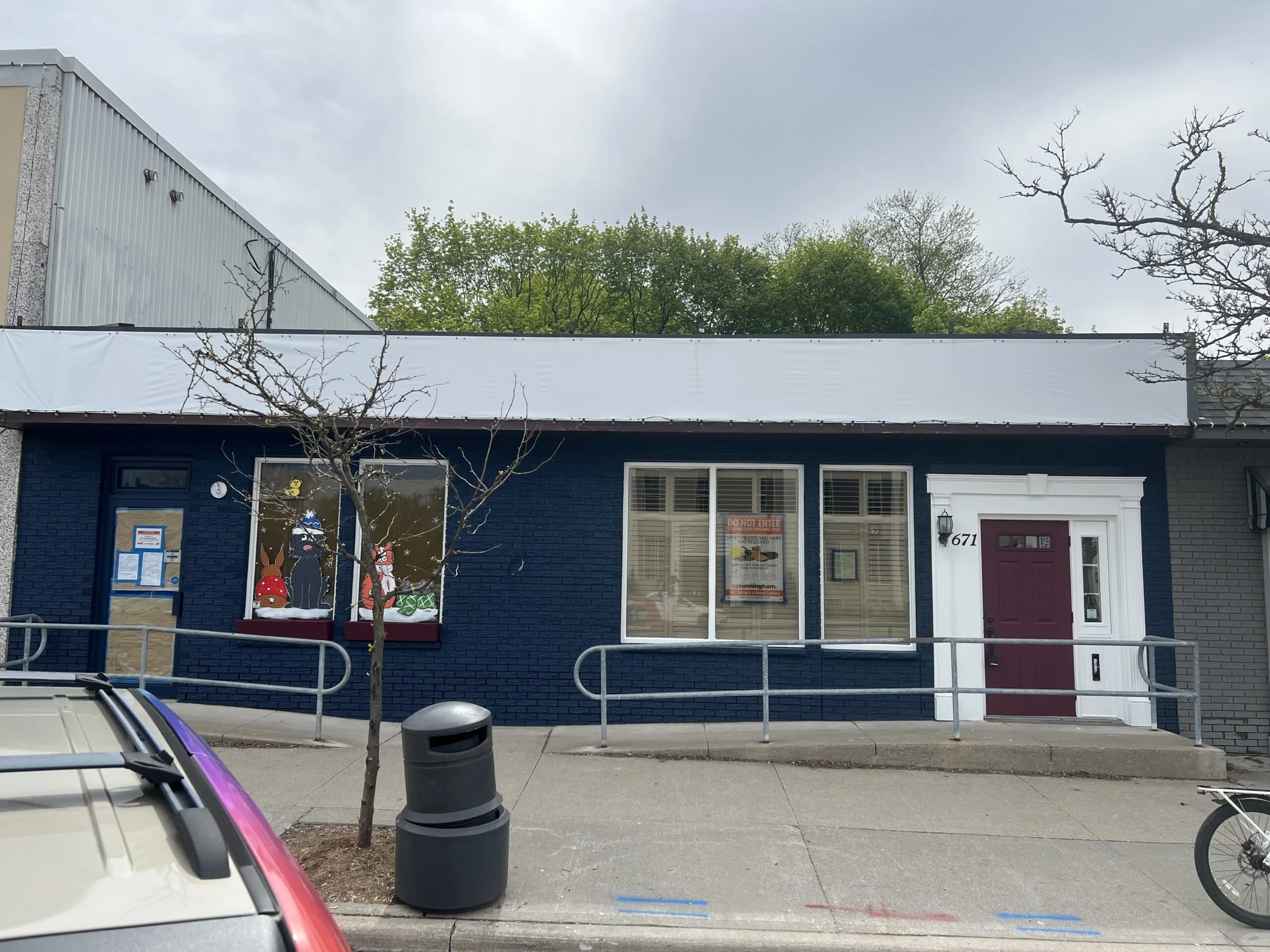 A blue brick building with a purple front door, four windows, some Christmas decorations in the window, a small tree in front, a trash can, a parked car partially visible, and a bicycle on the sidewalk.