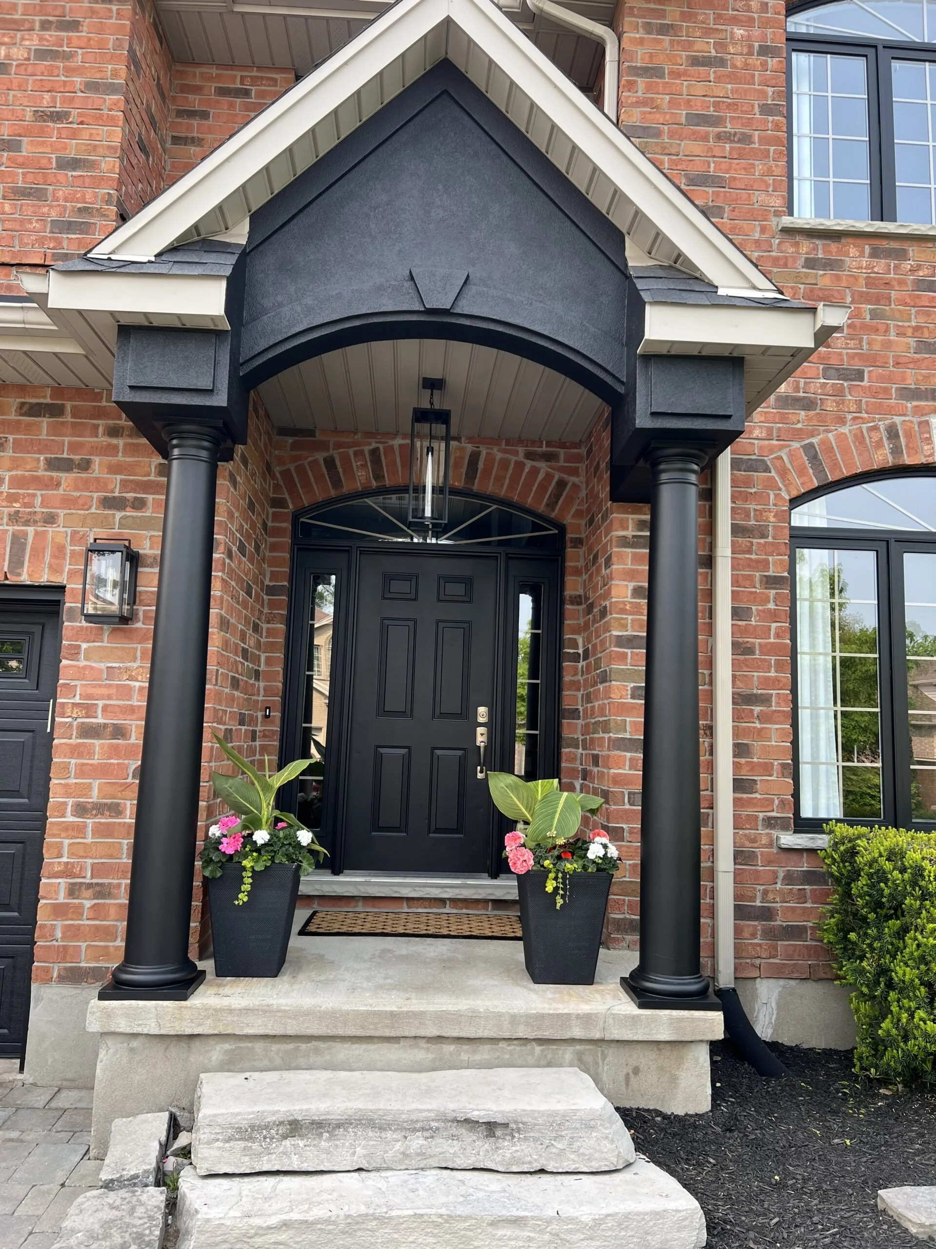 Front entrance of a brick house with black columns, black front door, two black planters with pink and white flowers, concrete steps, and a porch canopy.