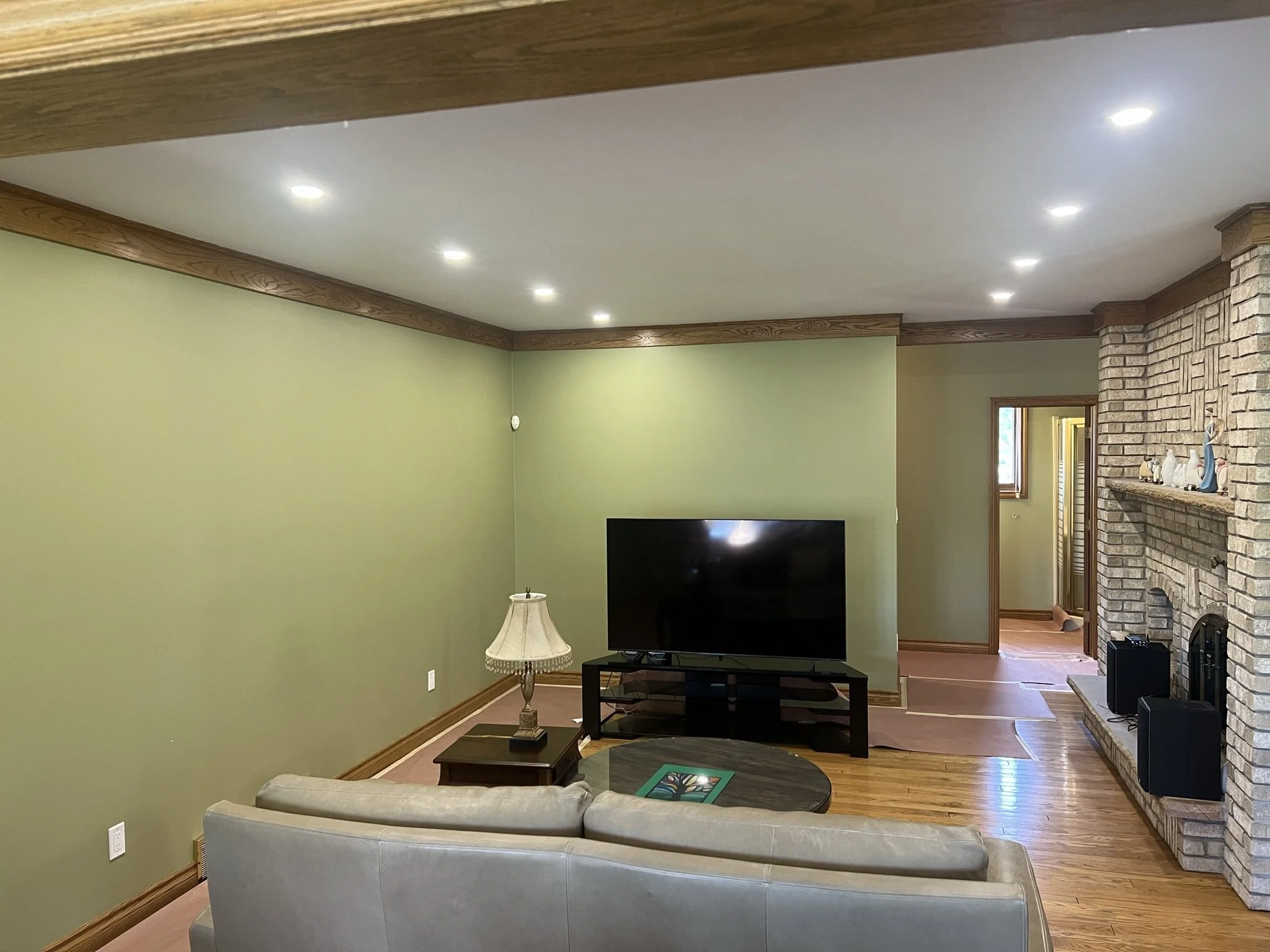 Living room with green walls, wooden trim, a cream sofa, a small wooden side table with a lamp, a TV on a stand, a round coffee table, a brick fireplace with decorations on the mantel, hardwood flooring, and a small window in the background.