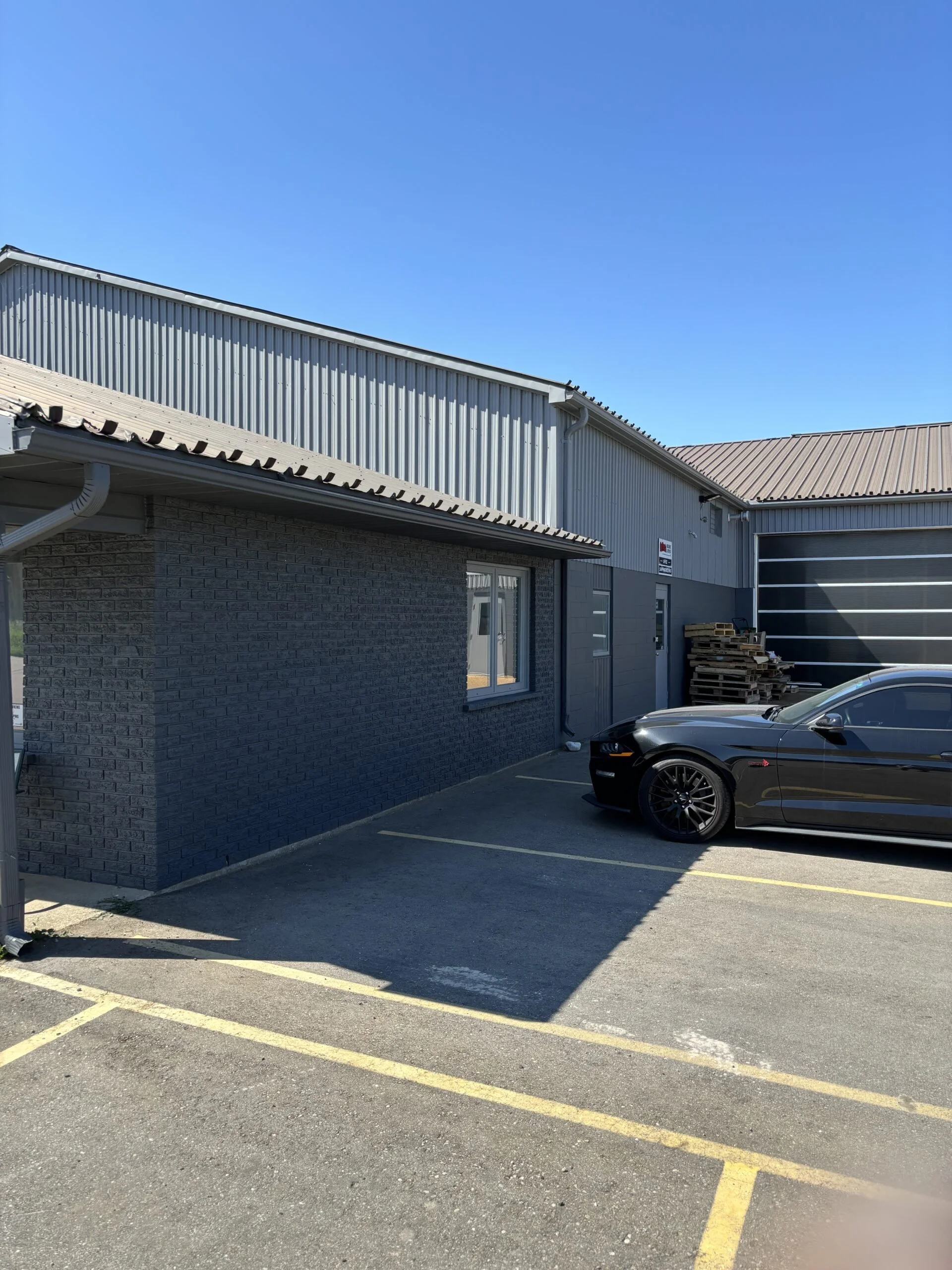A black sports car parked in a parking lot next to a gray brick building with a window. The building has a metal exterior with a corrugated roof and a stack of wooden pallets beside it. The sky is clear and blue.