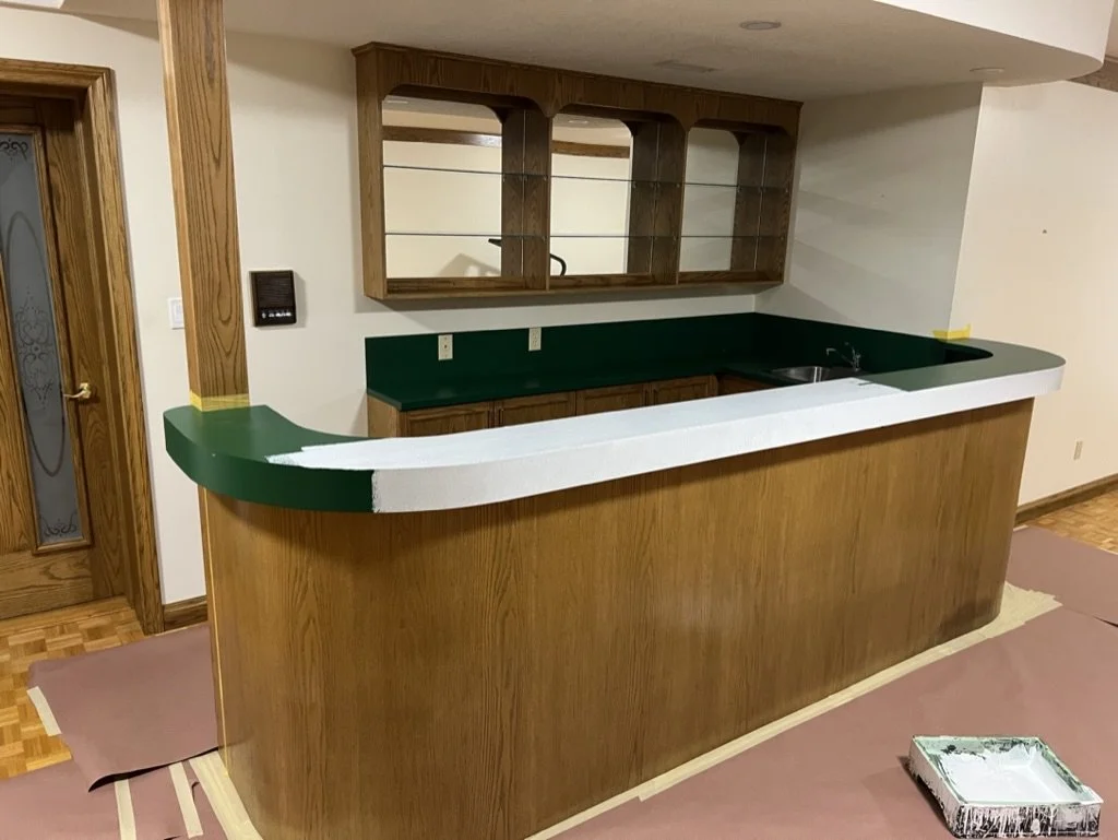 Kitchen with a wooden counter, green and white painted edges, and glass-front cabinets on the wall. There is a sink in the countertop and a door with frosted glass and decorative design on the left. A paint tray with white paint is on the floor.