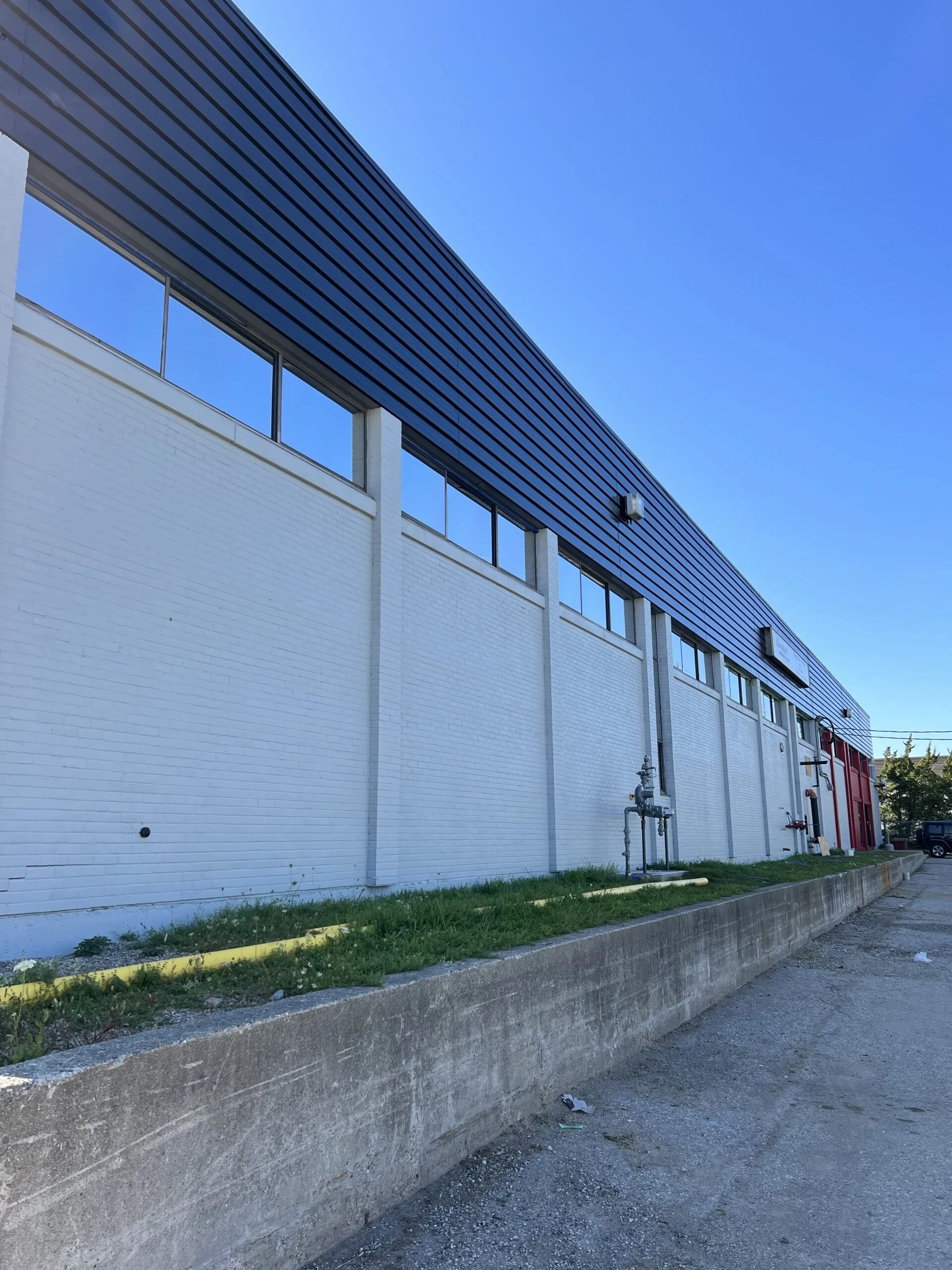 Exterior view of a large commercial or industrial building with a white brick lower wall, black metal siding upper wall, and several windows along the top. The area in front has a concrete curb and some grassy patches, with a few pipes and utility fi