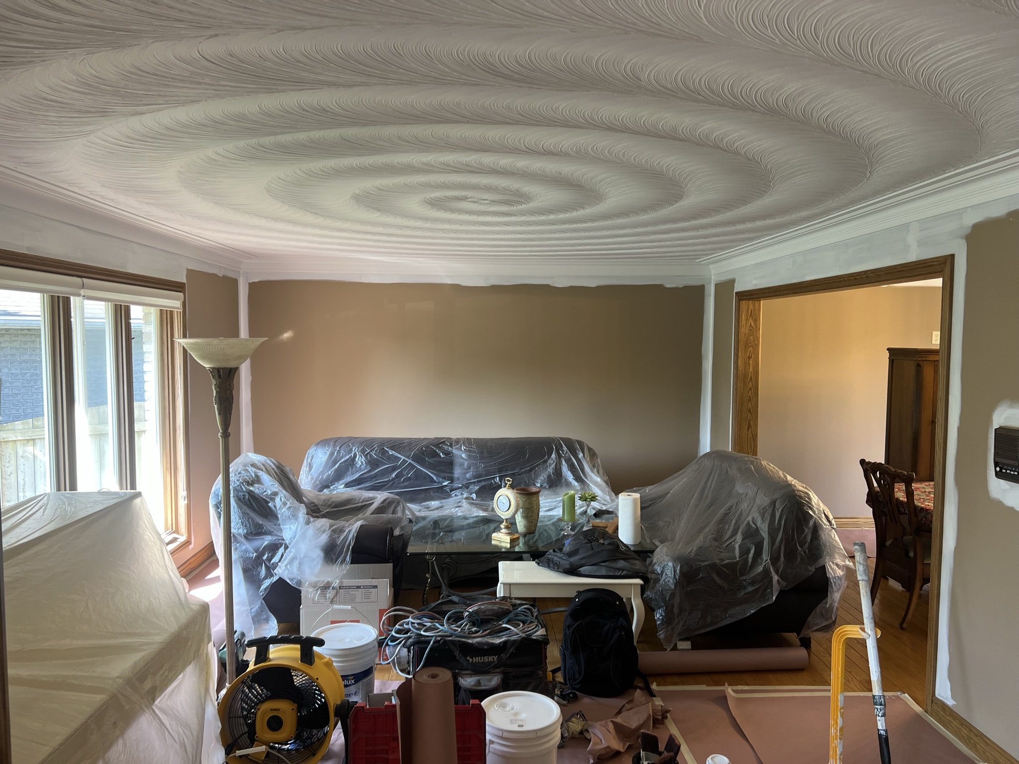 Living room under renovation with furniture covered in plastic, painting supplies, and tools scattered around.