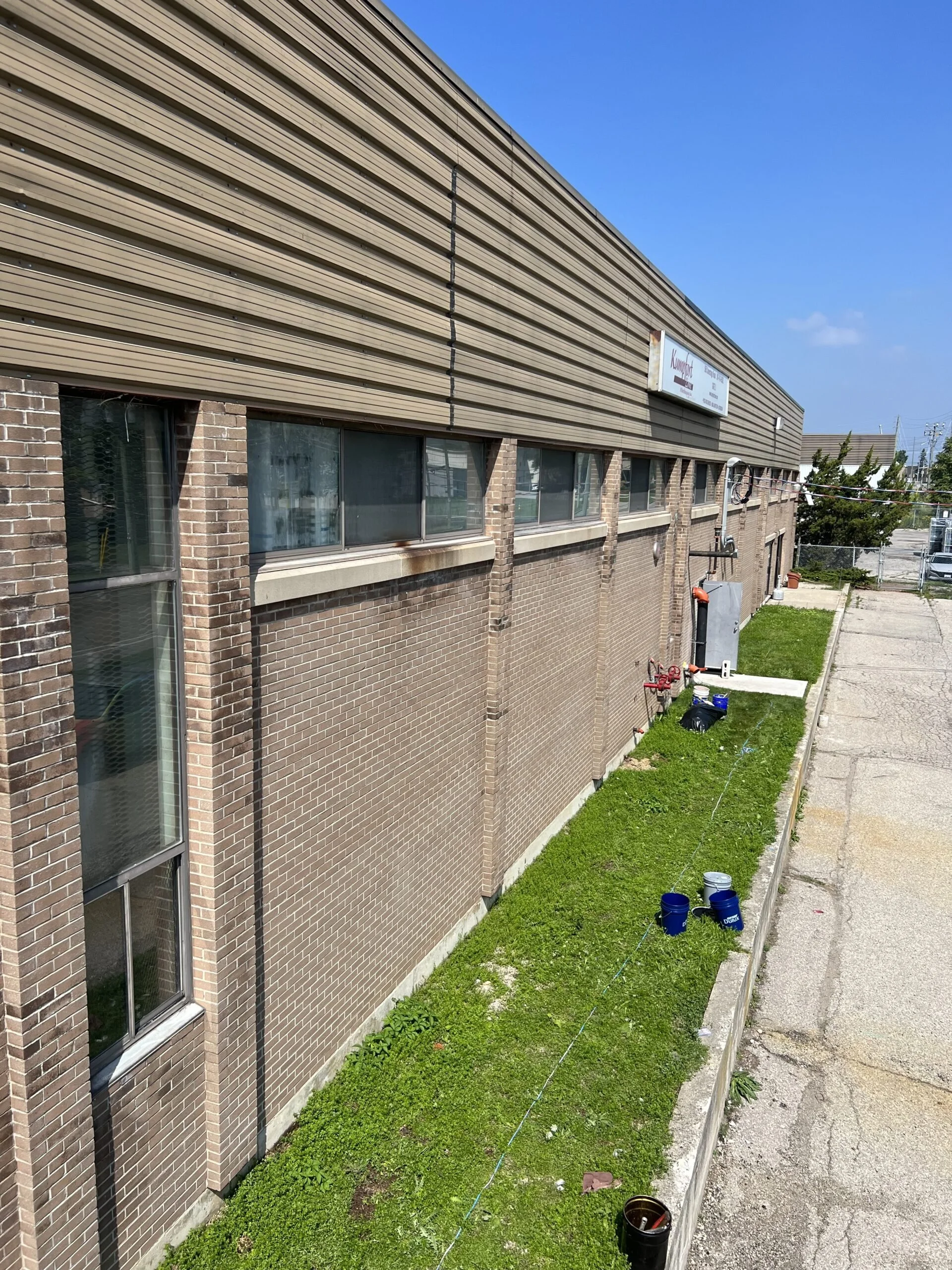 Side of a brick building with several windows, a signboard on the upper part, and a small grassy area with painting supplies, buckets, and a string line