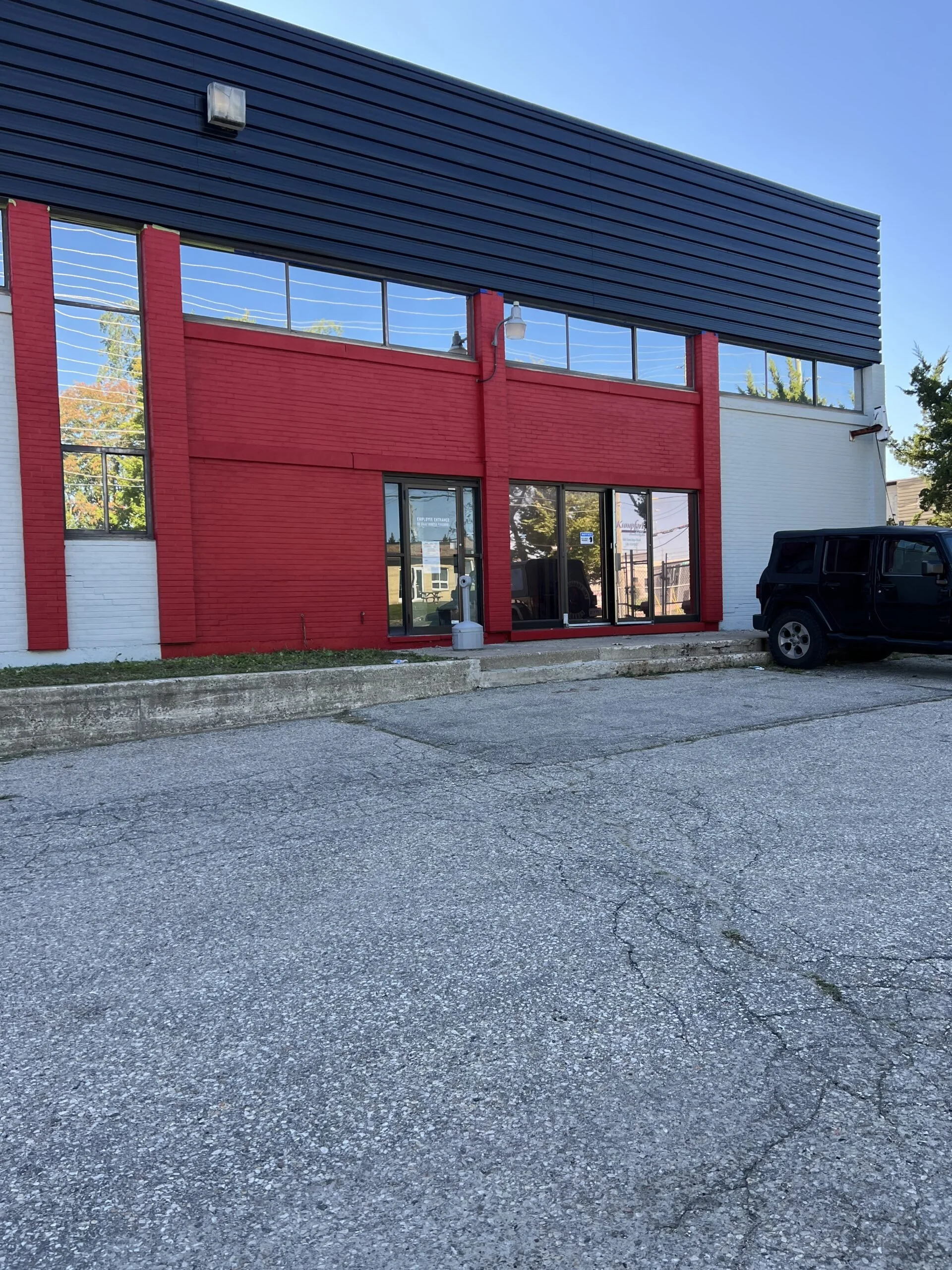 Front view of a modern commercial building with red and white exterior walls, large glass windows, and a black upper section, with a black vehicle parked in front on a cracked asphalt lot.