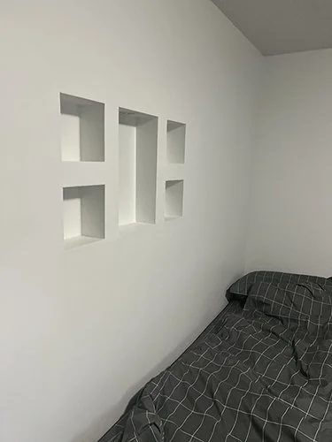 White wall with four square recessed shelves next to a bed with dark gray plaid bedding.