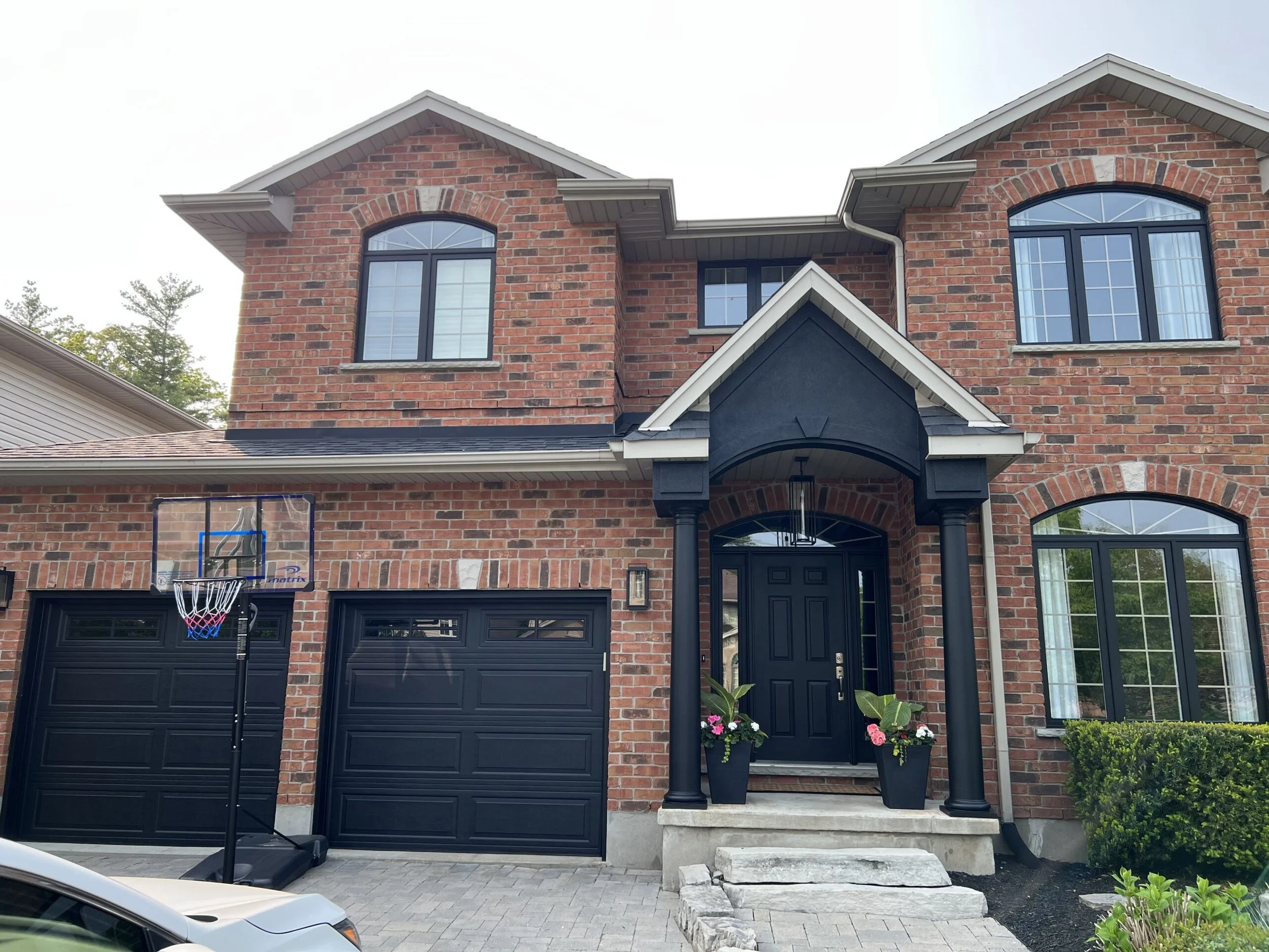 Front of a two-story brick house with black accents, a black front door, and a small porch with stairs. There are large windows with black frames and a basketball hoop near the garage.