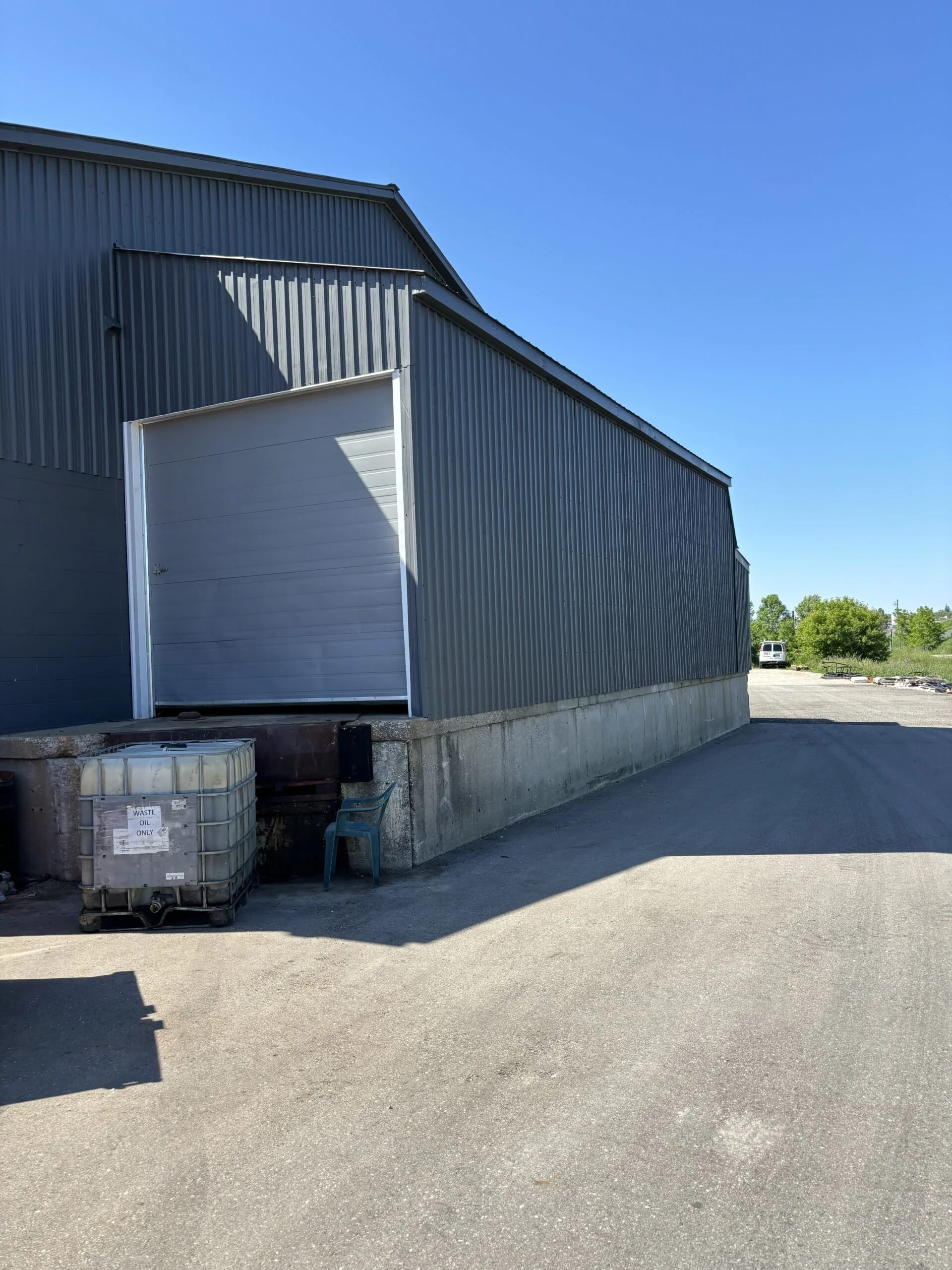 Side view of a large industrial metal building with a white roll-up door, a waste oil container, a blue plastic chair, and an open paved area under a clear blue sky.