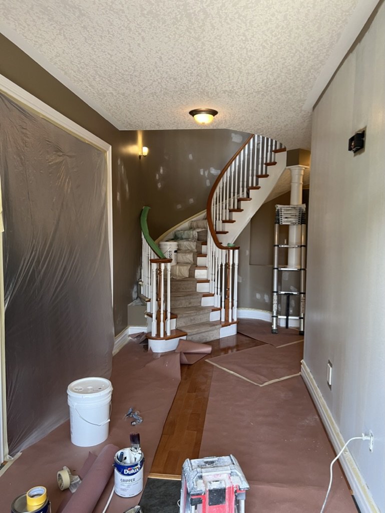 Interior of a house under renovation with a curved staircase, partially covered with protection plastic, and painting supplies on the floor.