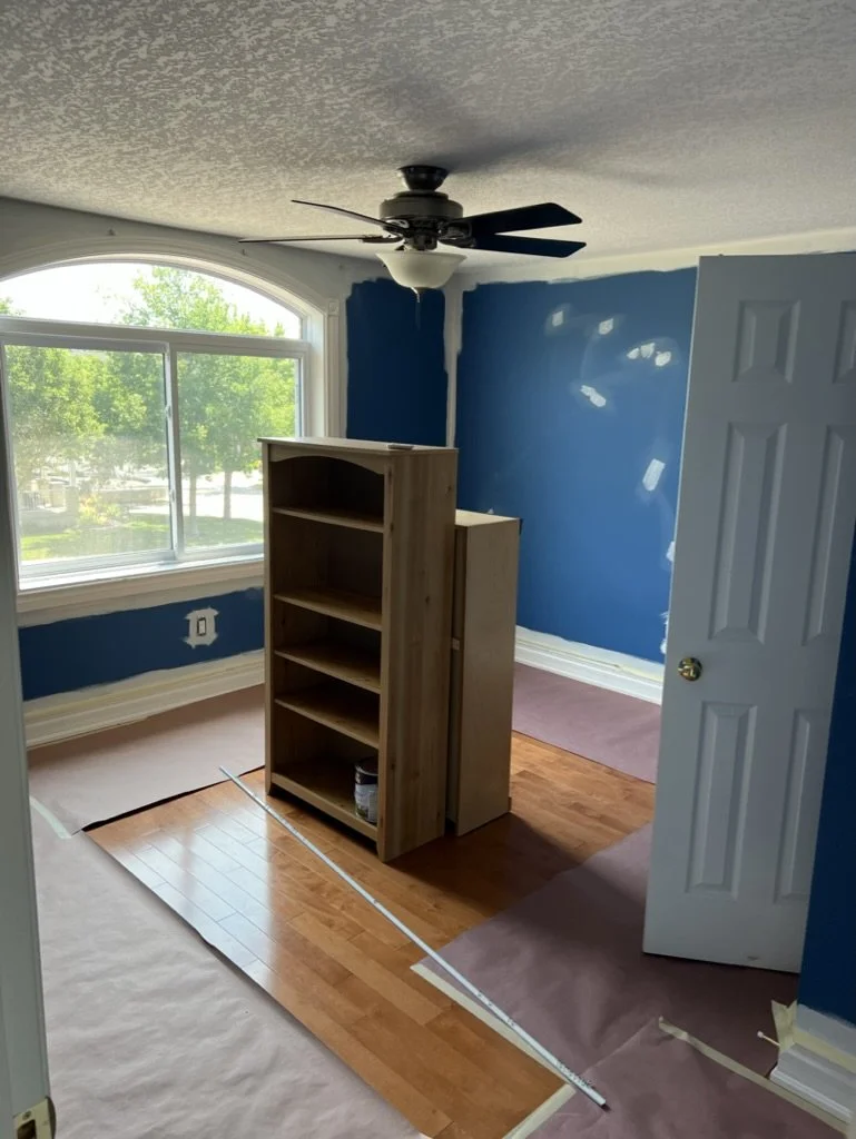 Room under renovation with blue walls, wooden floor partially covered with protective paper, a wooden shelf in the center, a large window with greenery outside, and a ceiling fan.