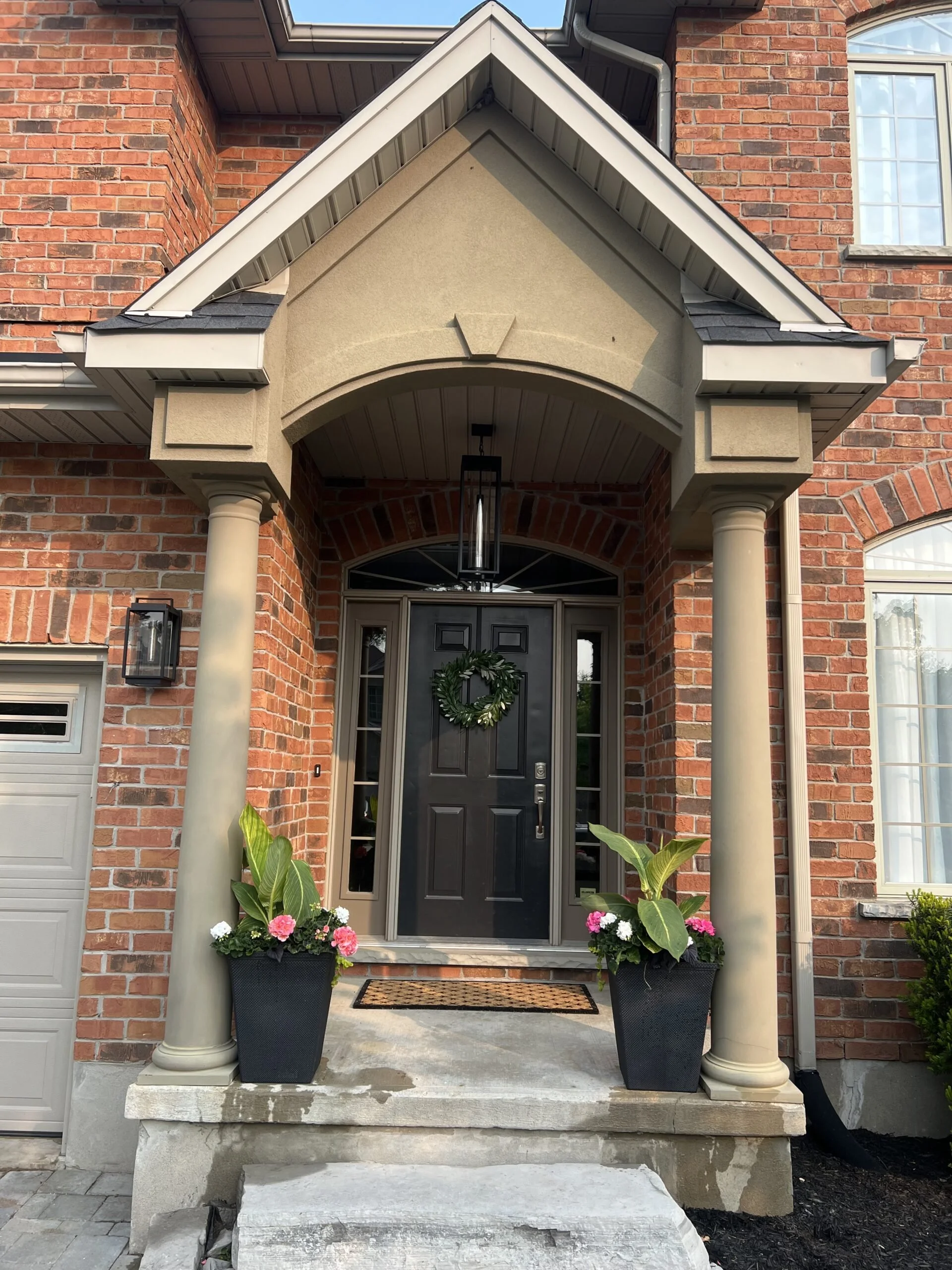 Front porch of a brick house with black door decorated with a green wreath, two large flower pots with pink and white flowers and green leaves, and a doormat, with stairs and columns supporting a small roof over the entrance.