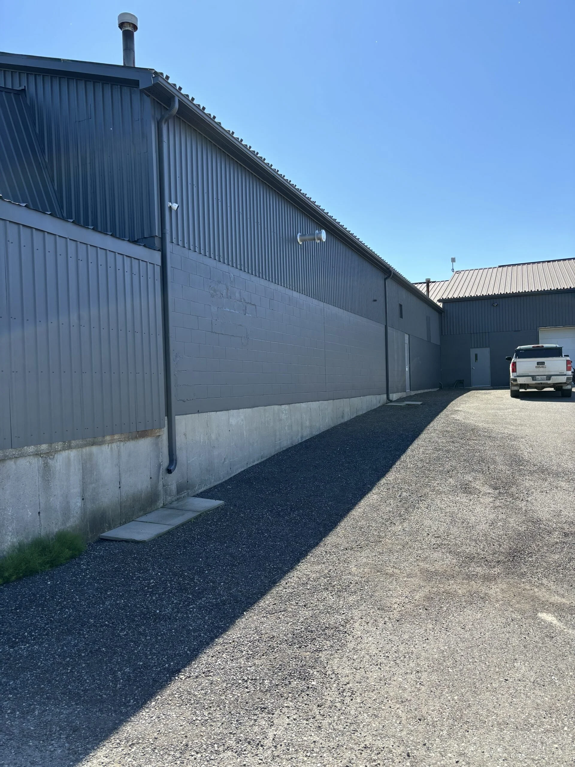 An industrial building with metal siding, concrete foundation, and a parking area with a white pickup truck. Clear blue sky and shadows from the building are visible.