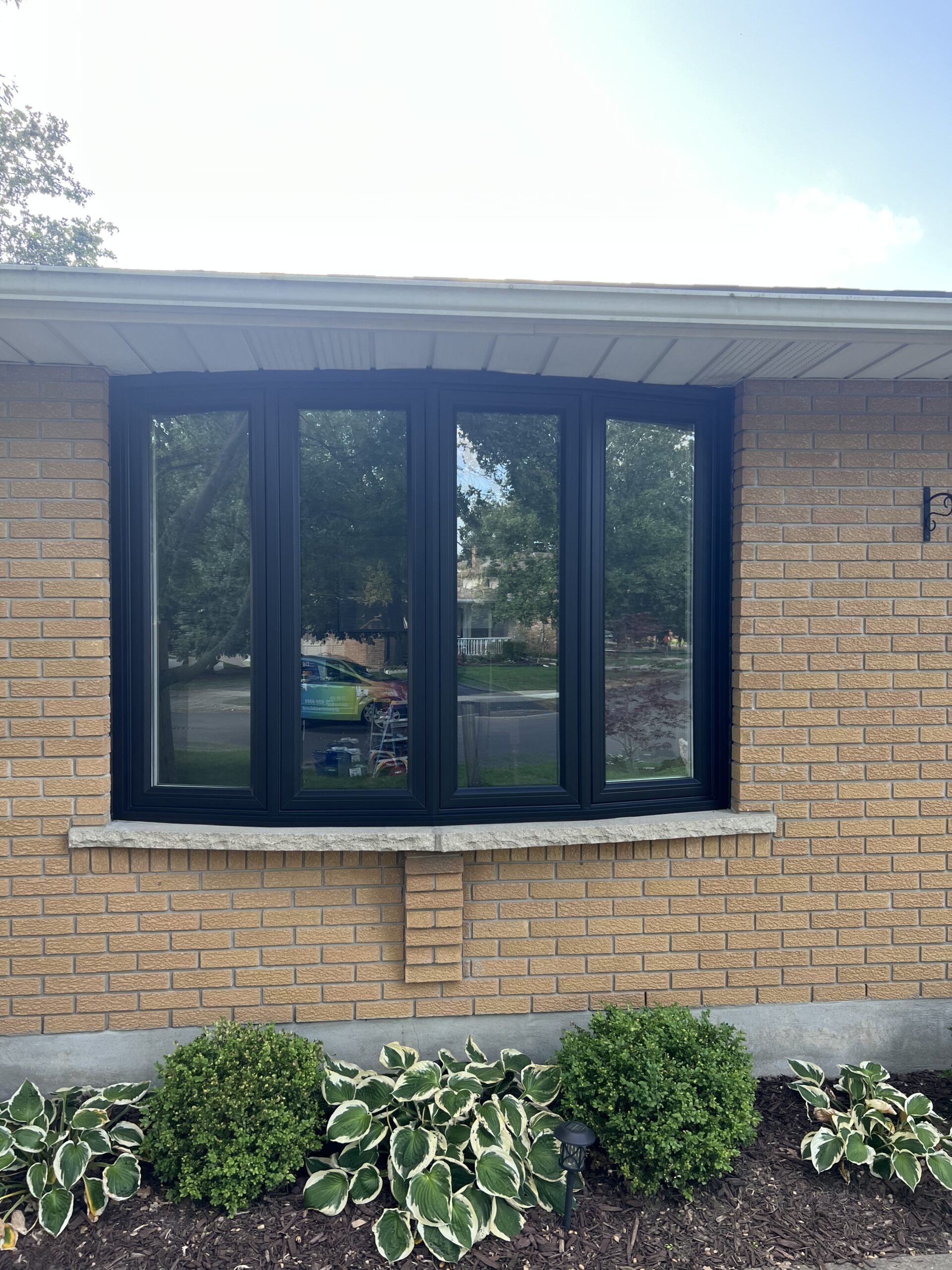 A brick house with a large, black-framed bay window and a flower bed with green shrubs and variegated hostas in front.